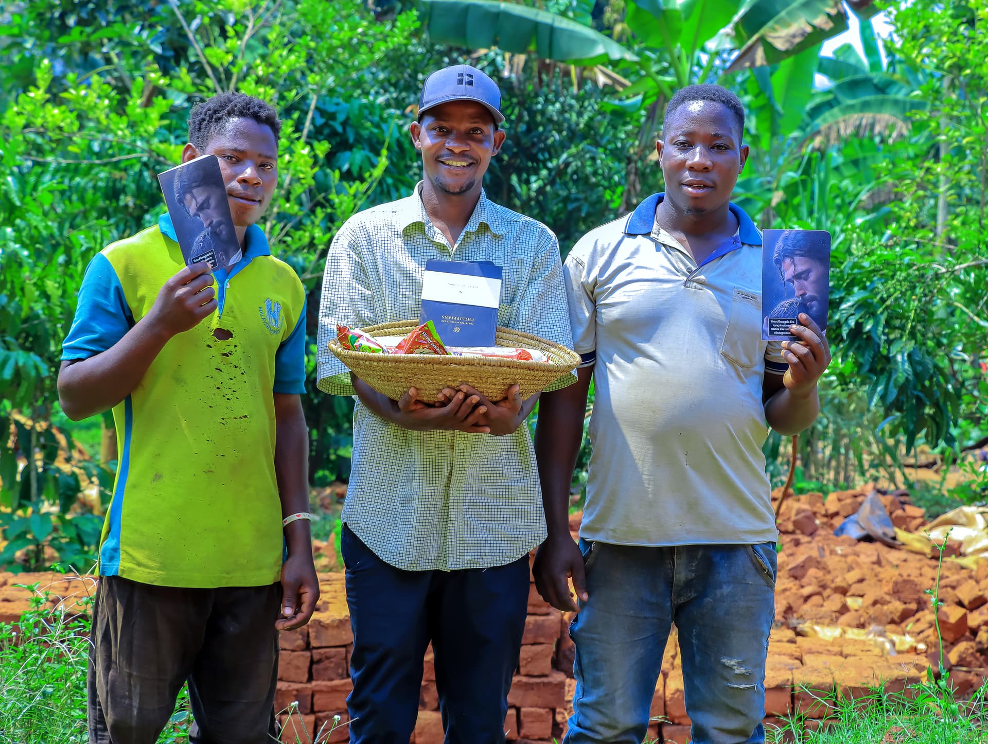 Three men stand together outdoors, one holding a woven plate of gospel tracts.