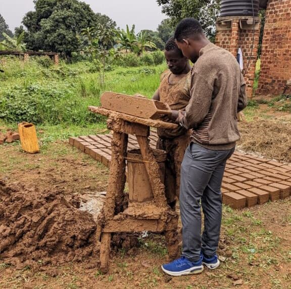 Two men mold wet clay into bricks using a wooden brick press near a pile of finished bricks.
