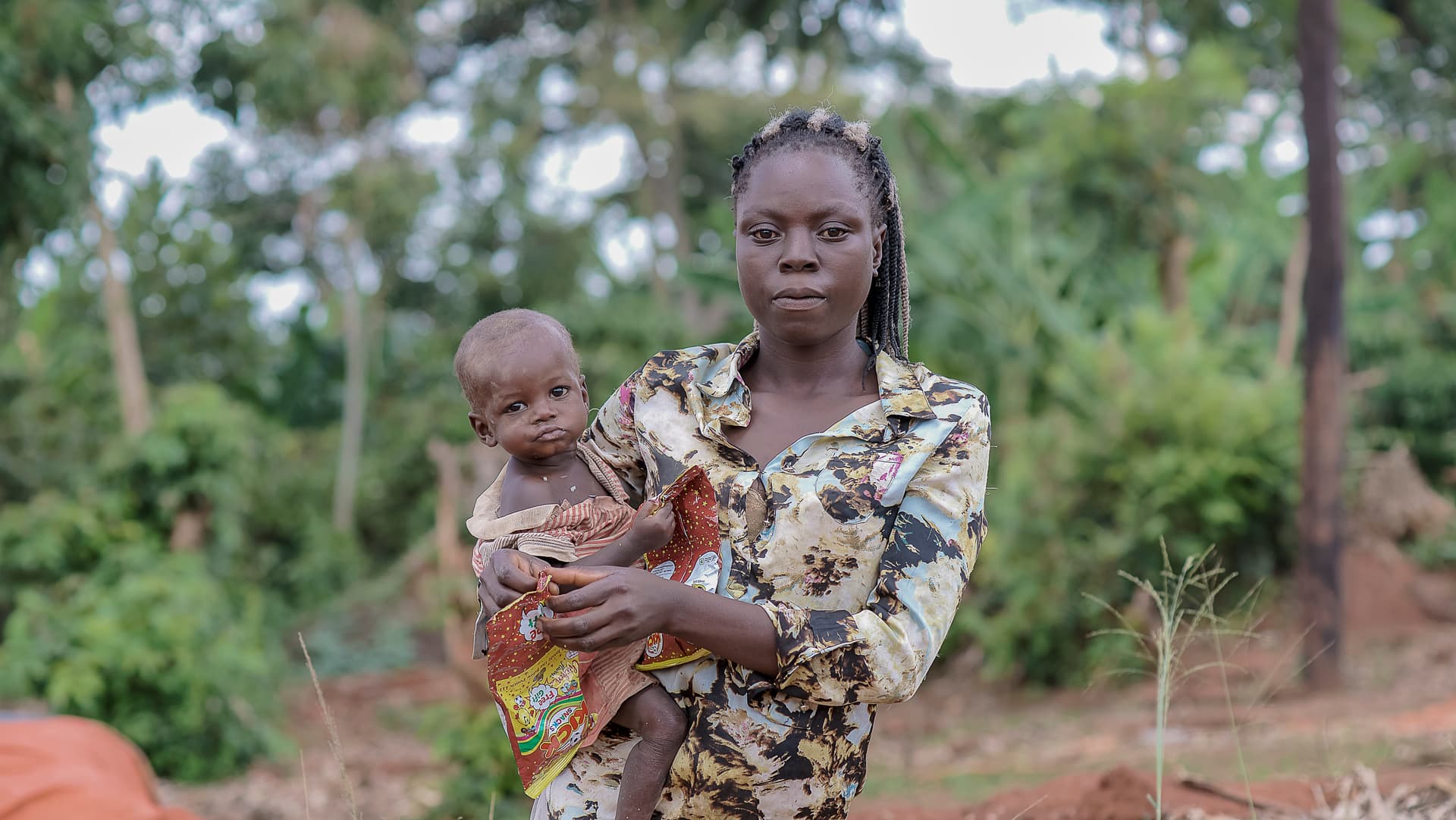 A woman holds an infant while standing outdoors in a wooded area with another woman nearby.