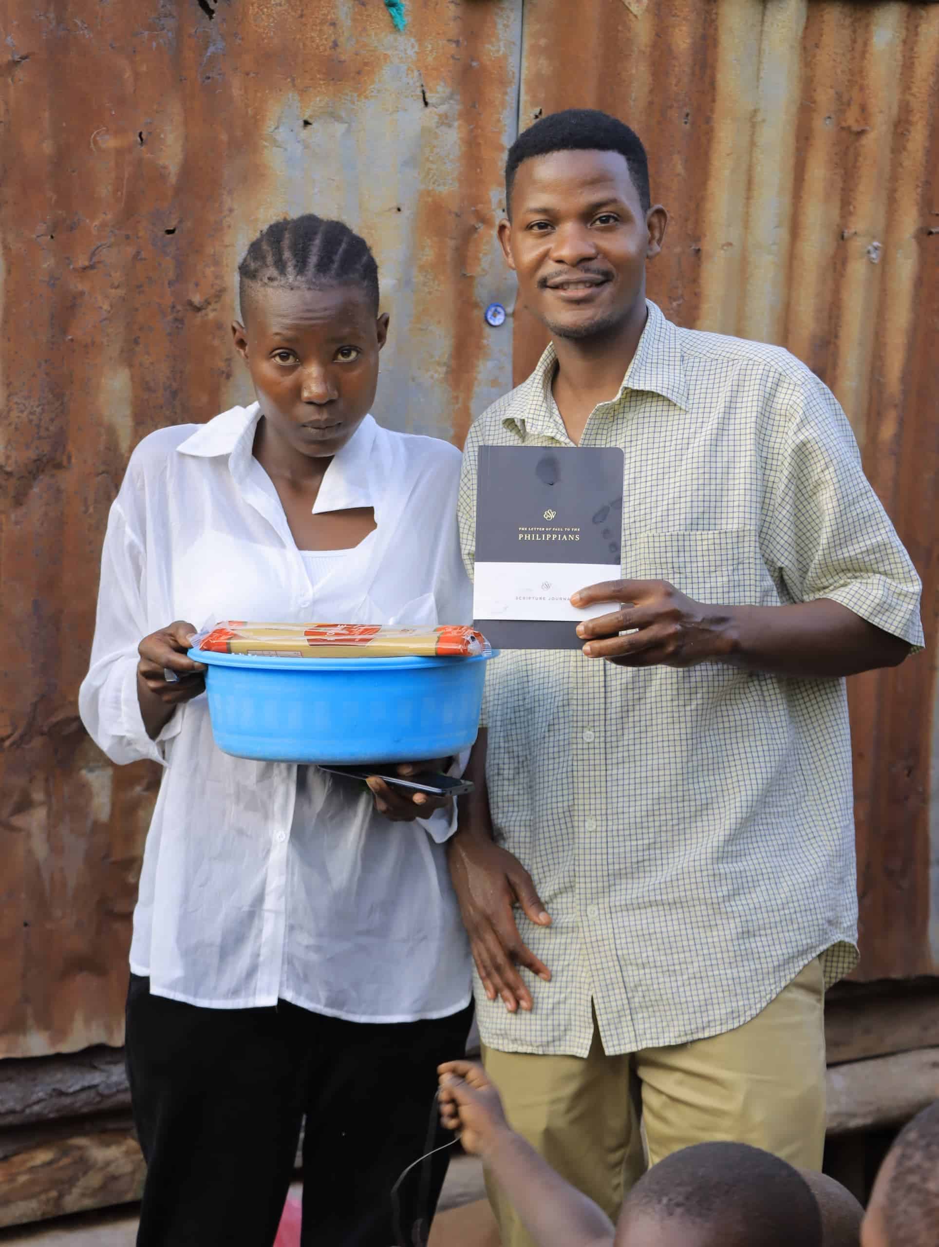 A young woman and a man stand together outdoors holding an orange basin of food gifts and a booklet.