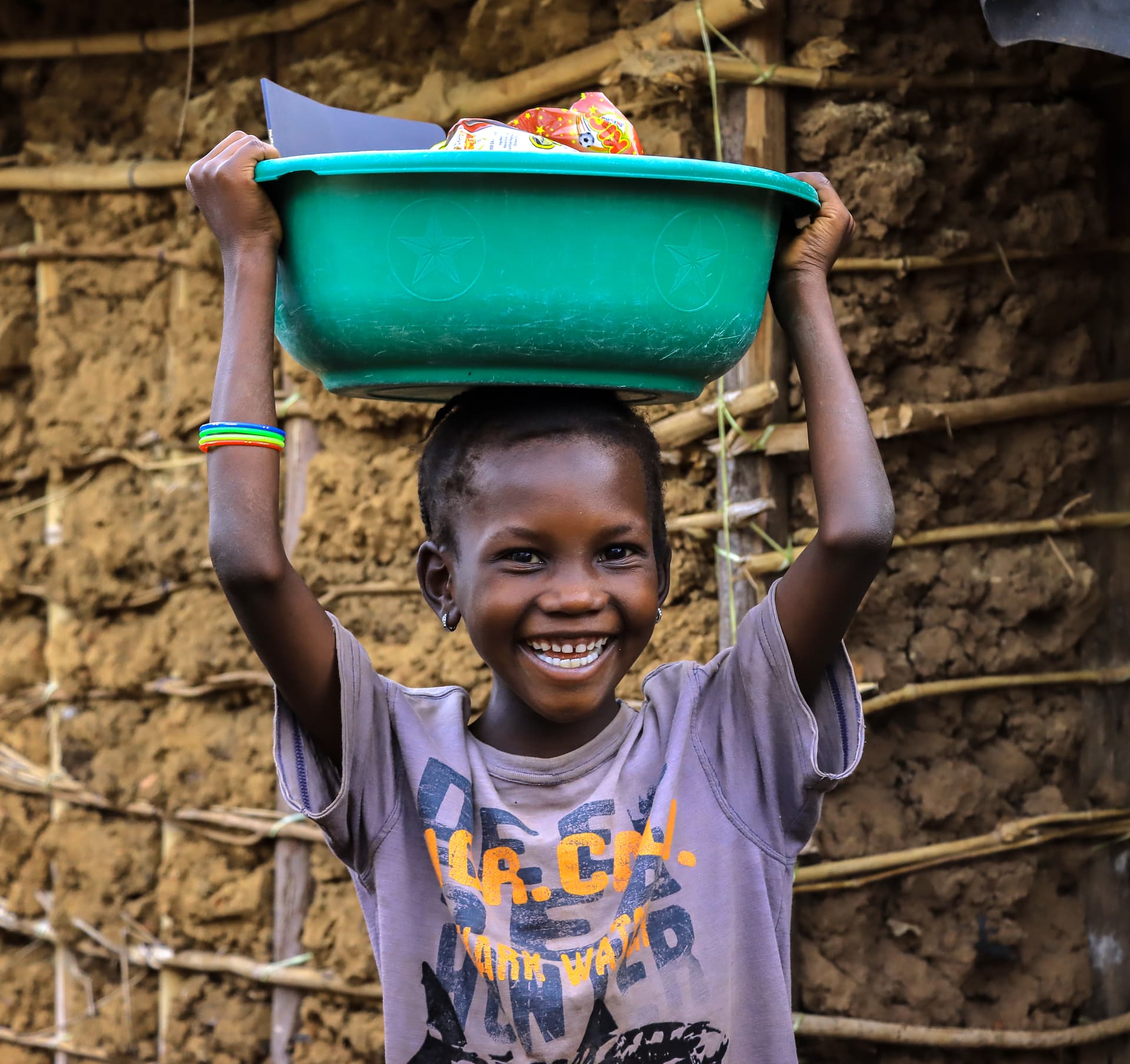 A smiling boy balances a green basin of food gifts on his head outside a mud-brick home.