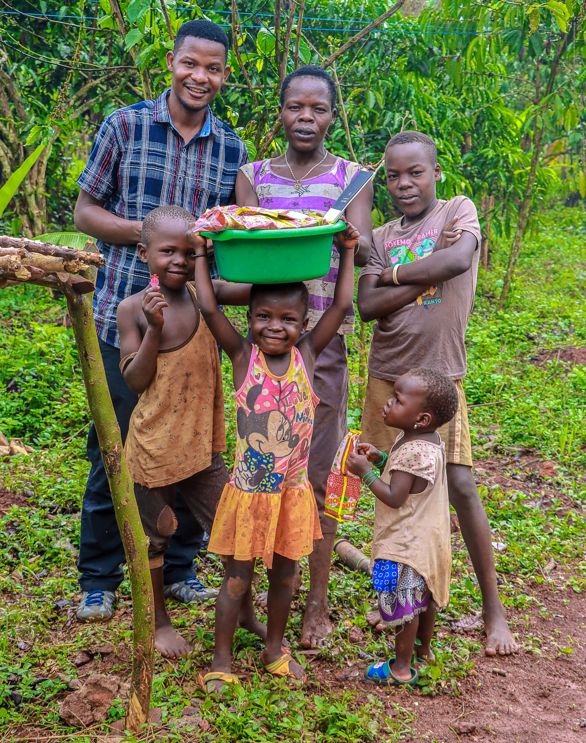 A man and three children stand together outdoors holding a green basin of food gifts by a wooden fence.