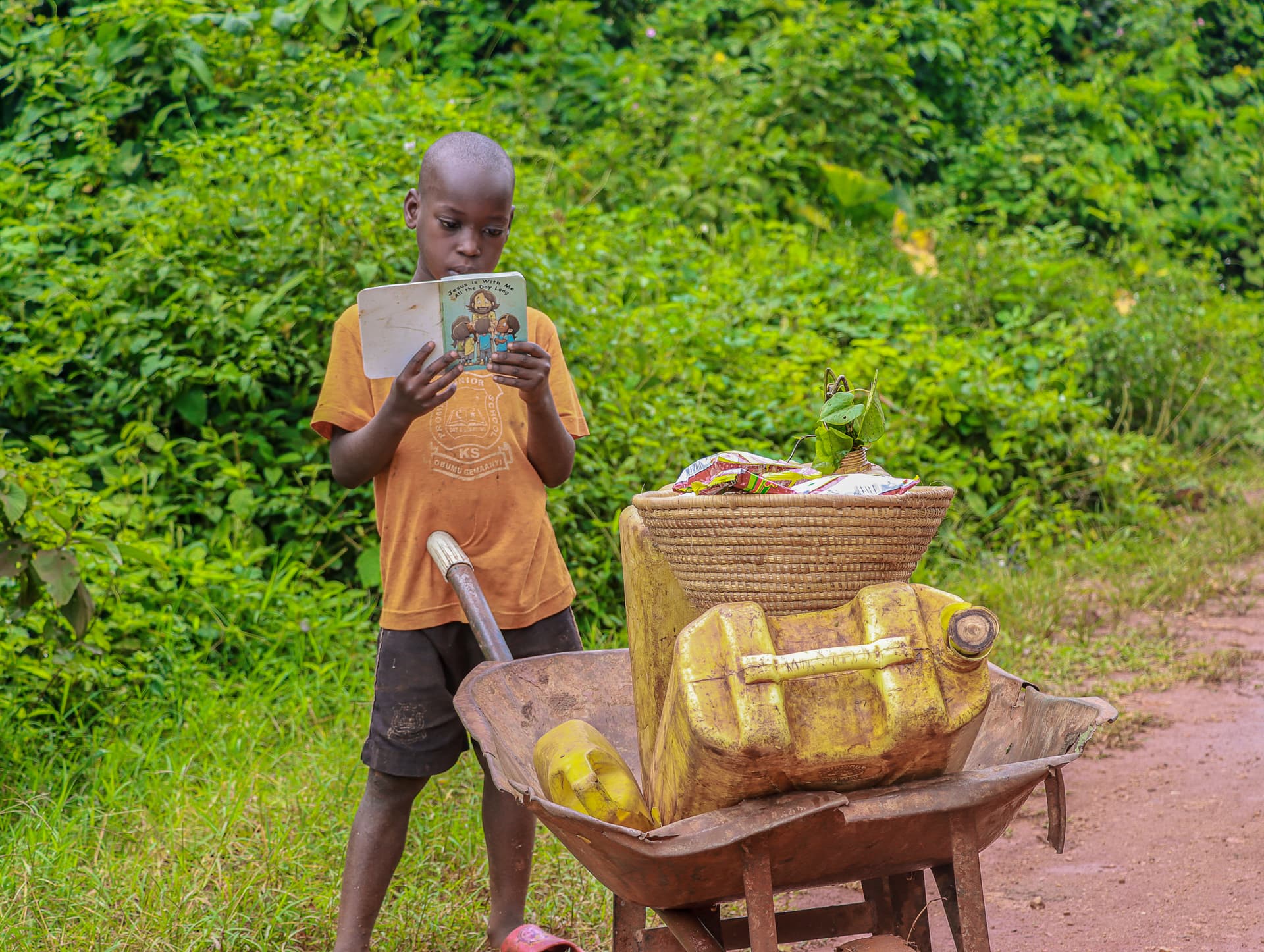 A boy reads a booklet beside a wooden wheelbarrow loaded with jerrycans and produce on a dirt path.