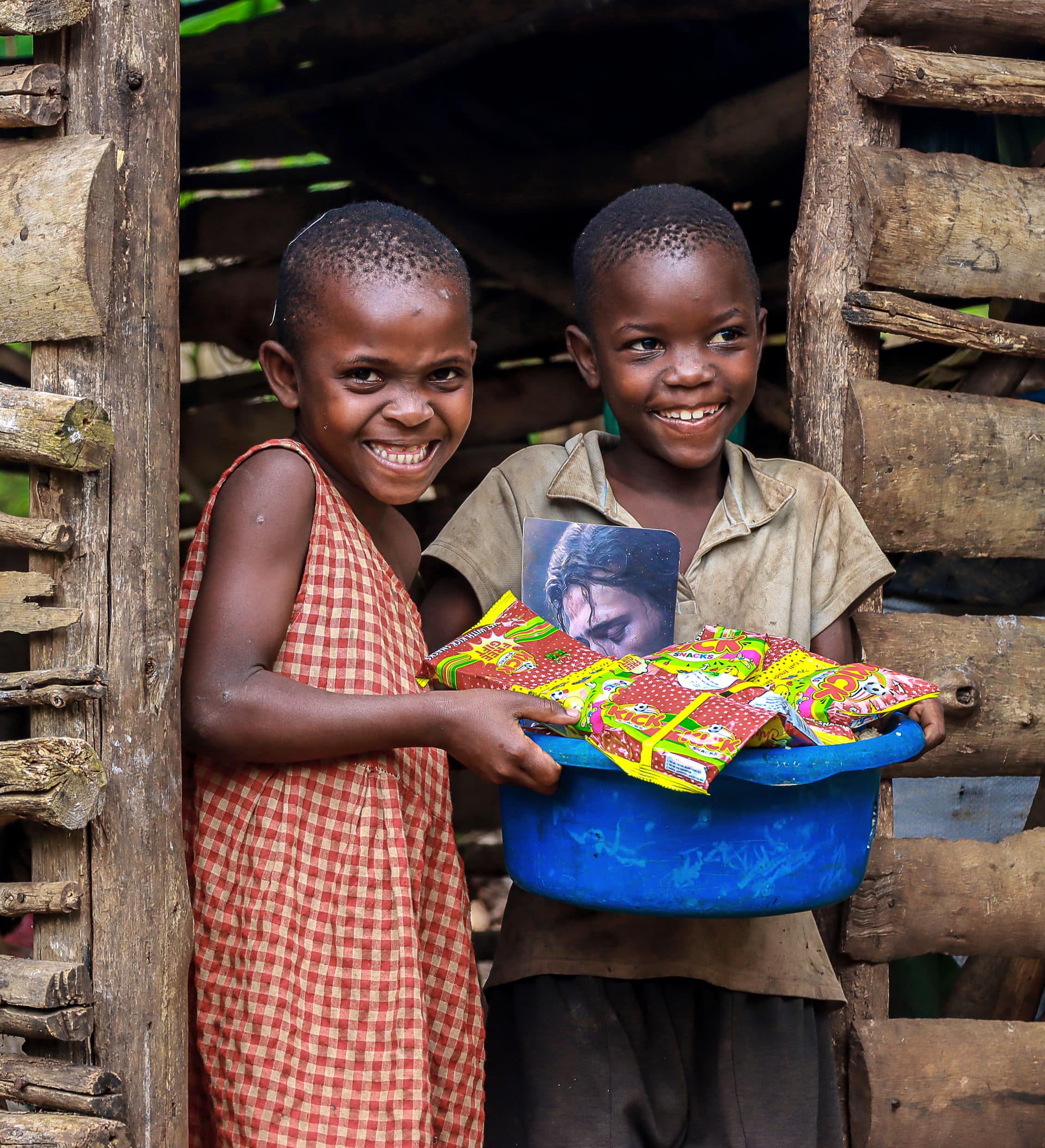 Two children hold a shared blue basin of food gift packages and a booklet beside a wooden structure.