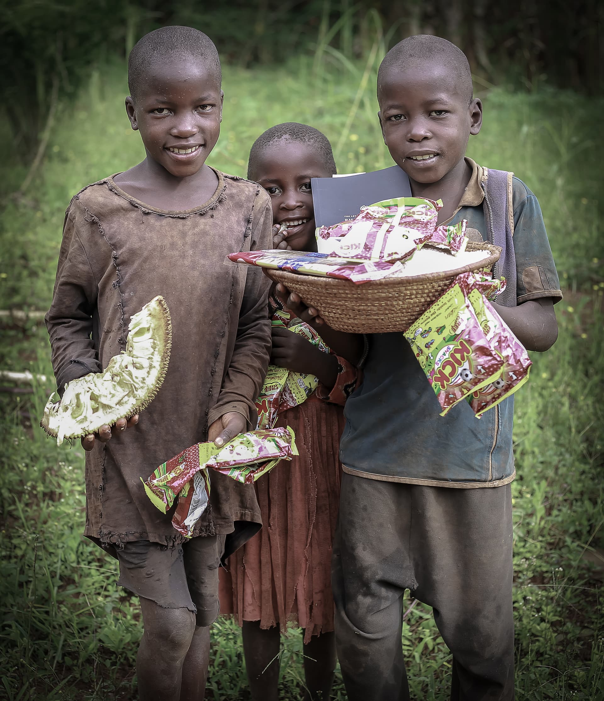 Three boys stand together outdoors holding food gift packages and a woven basket.