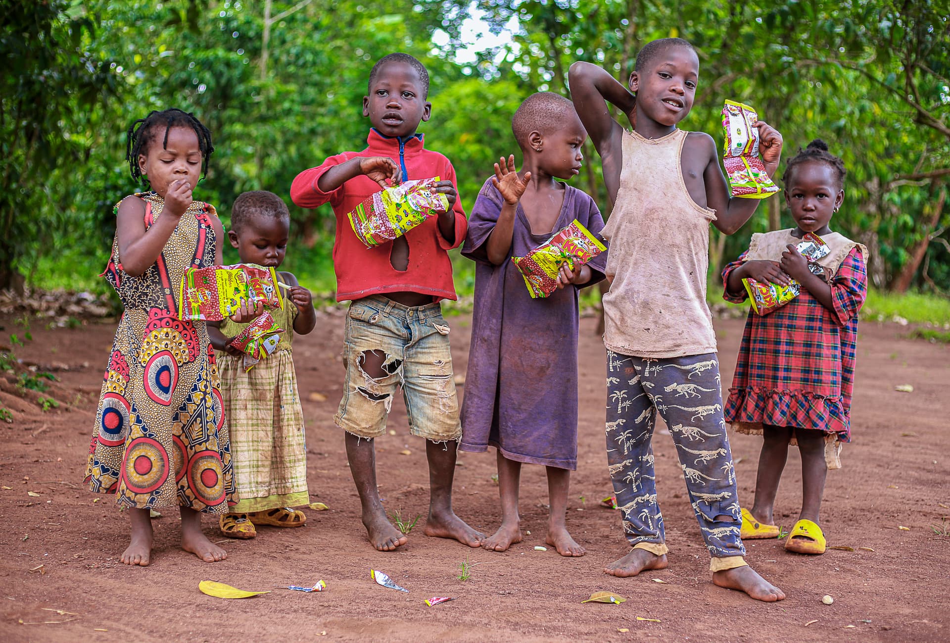 A group of five children stand together on a dirt road holding food gift packages.