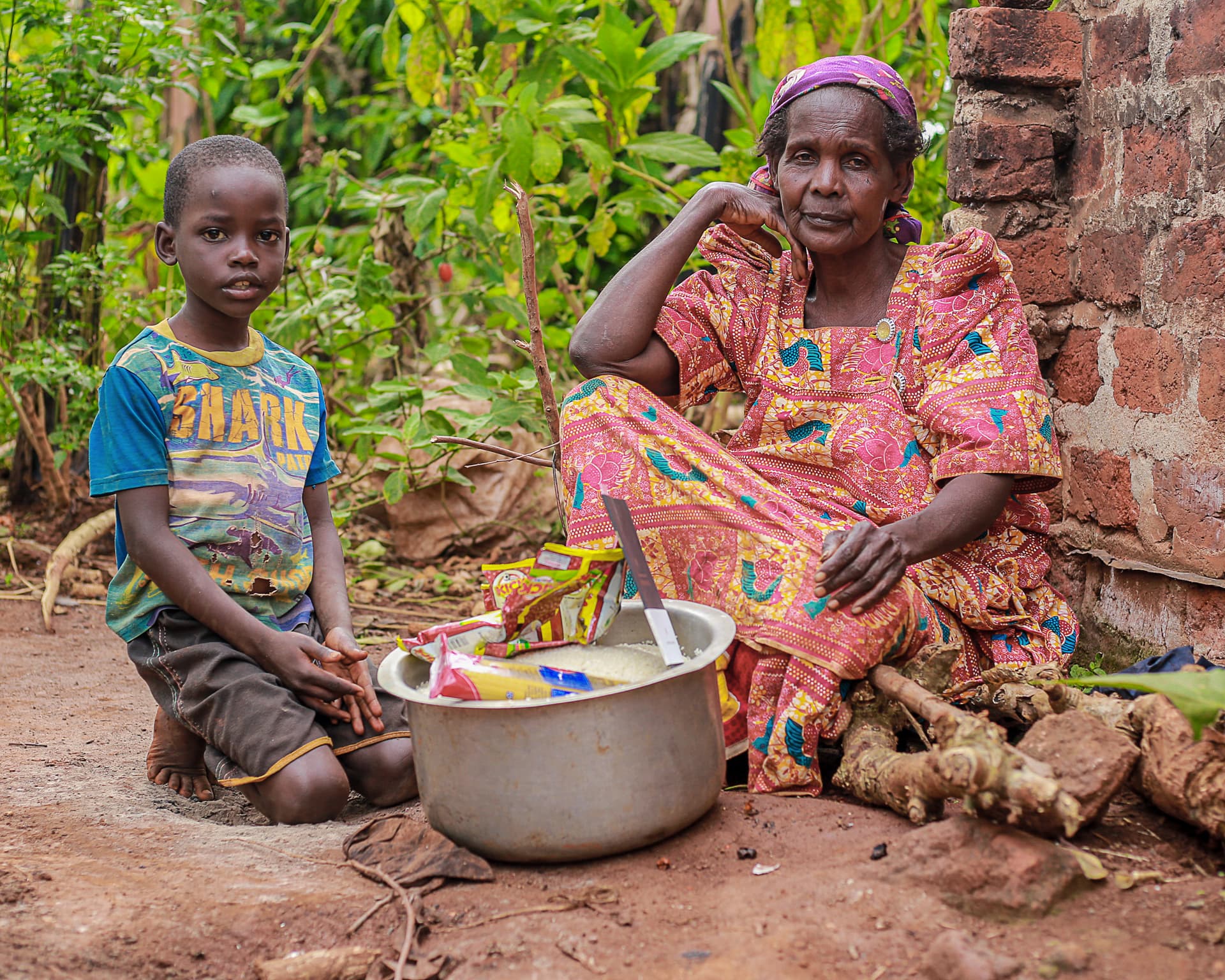 A boy sits beside an elderly woman outdoors with a large metal basin between them near a brick structure.