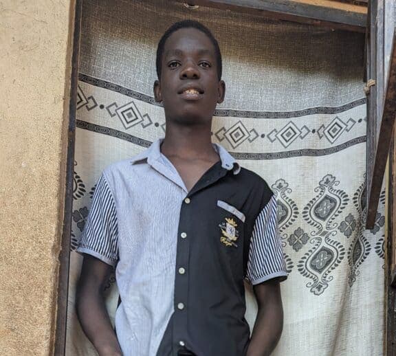 A teenage boy in a crested school uniform shirt stands indoors in front of a curtain, identified as Joseph.