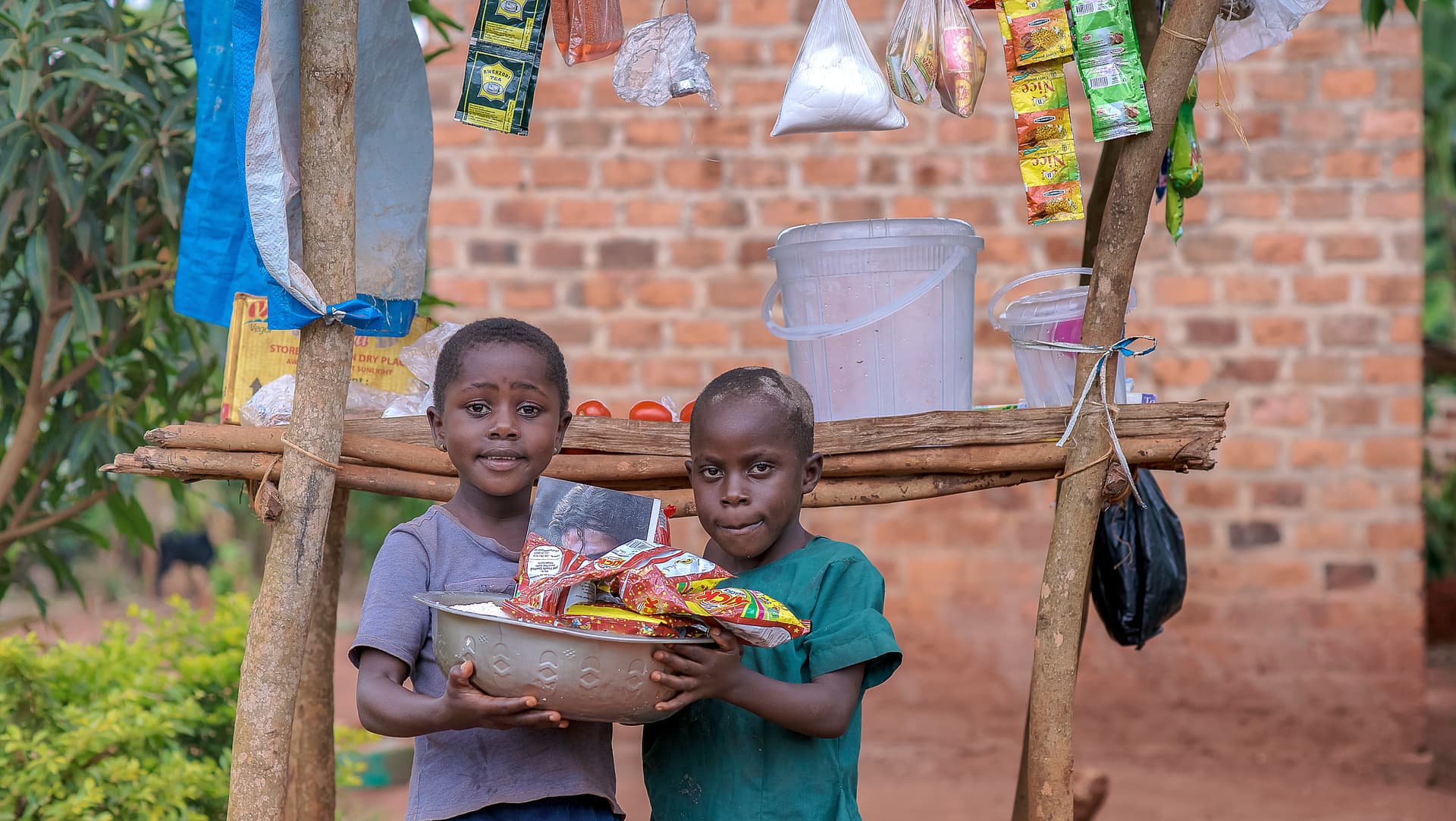 Two boys stand together outdoors holding a plate of food near a brick building with laundry hanging behind them.
