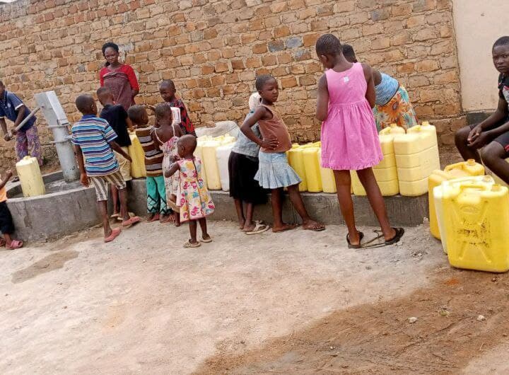 A line of children stands outdoors near a brick wall beside stacked yellow water jerrycans.