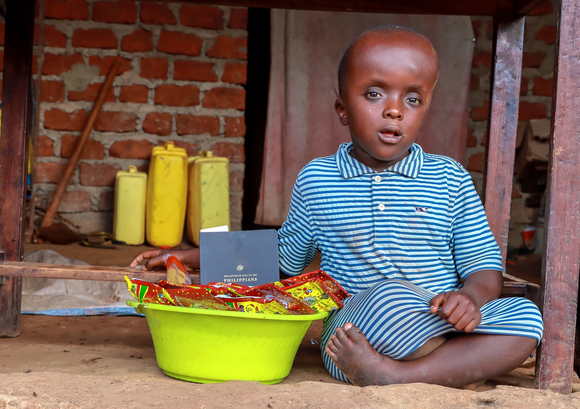 A boy in a striped shirt sits on the ground next to a green basin filled with food gift packages.