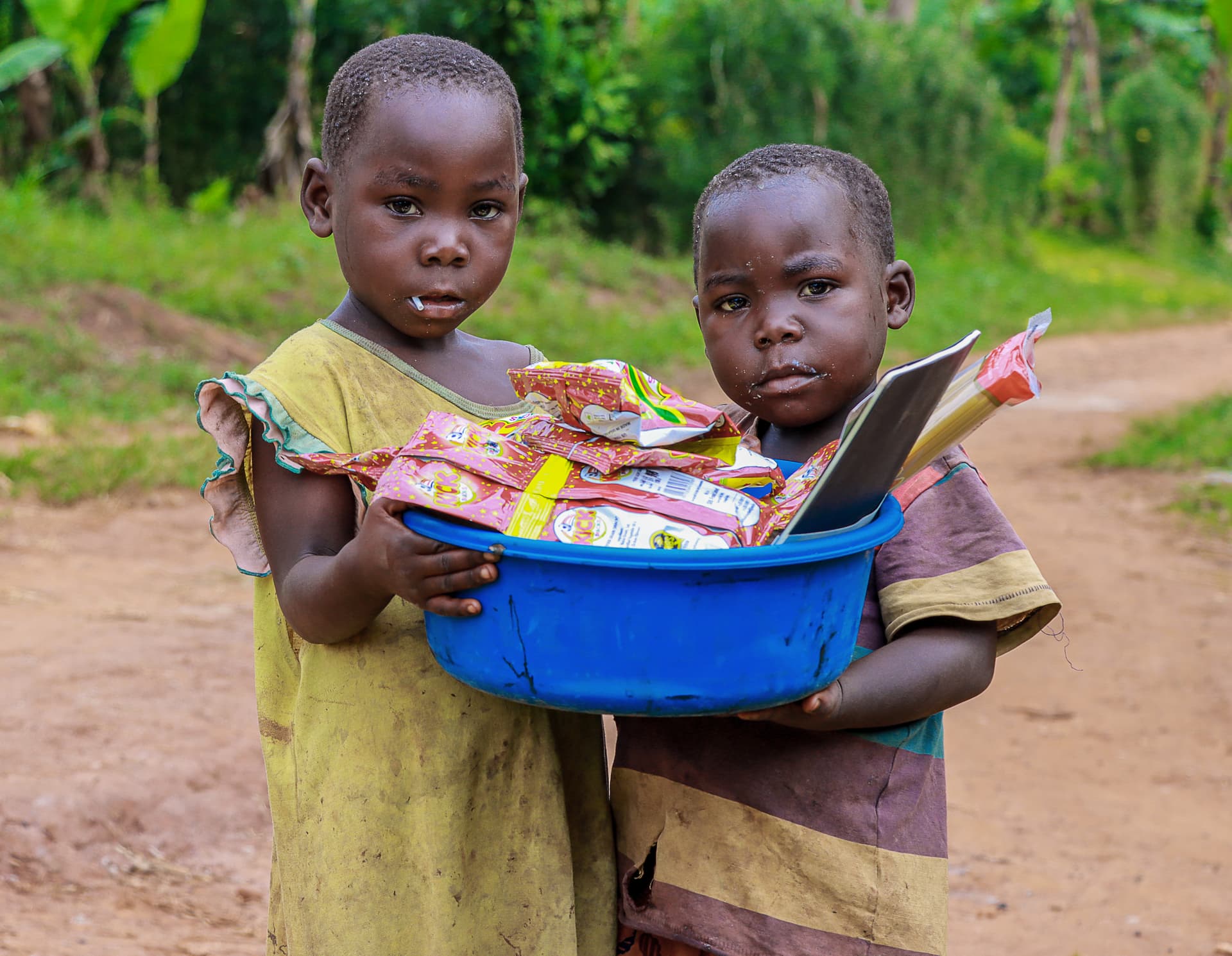 Two young Ugandan girls in yellow dresses hold a blue basin filled with JFM food gift packages on a dirt road.