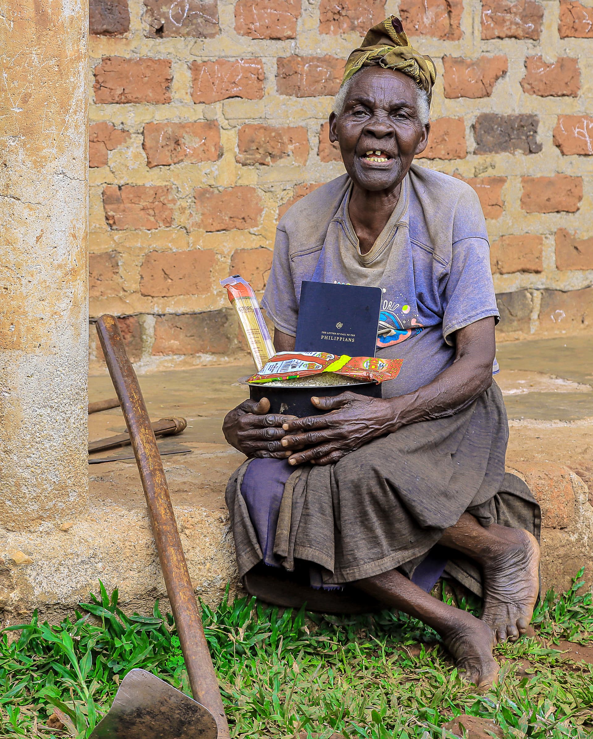 An elderly woman in a headscarf sits outside a brick house holding a food gift basin and booklet, walking stick beside her.