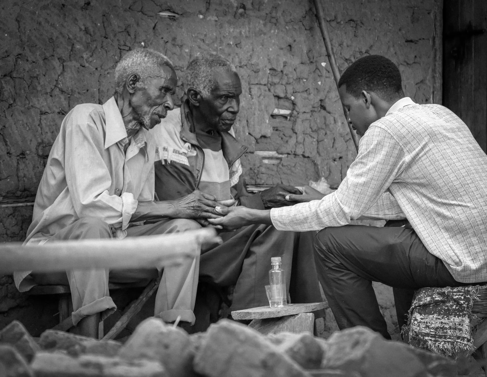 Black-and-white photo of two elderly men and a younger man seated at a table against a mud wall.