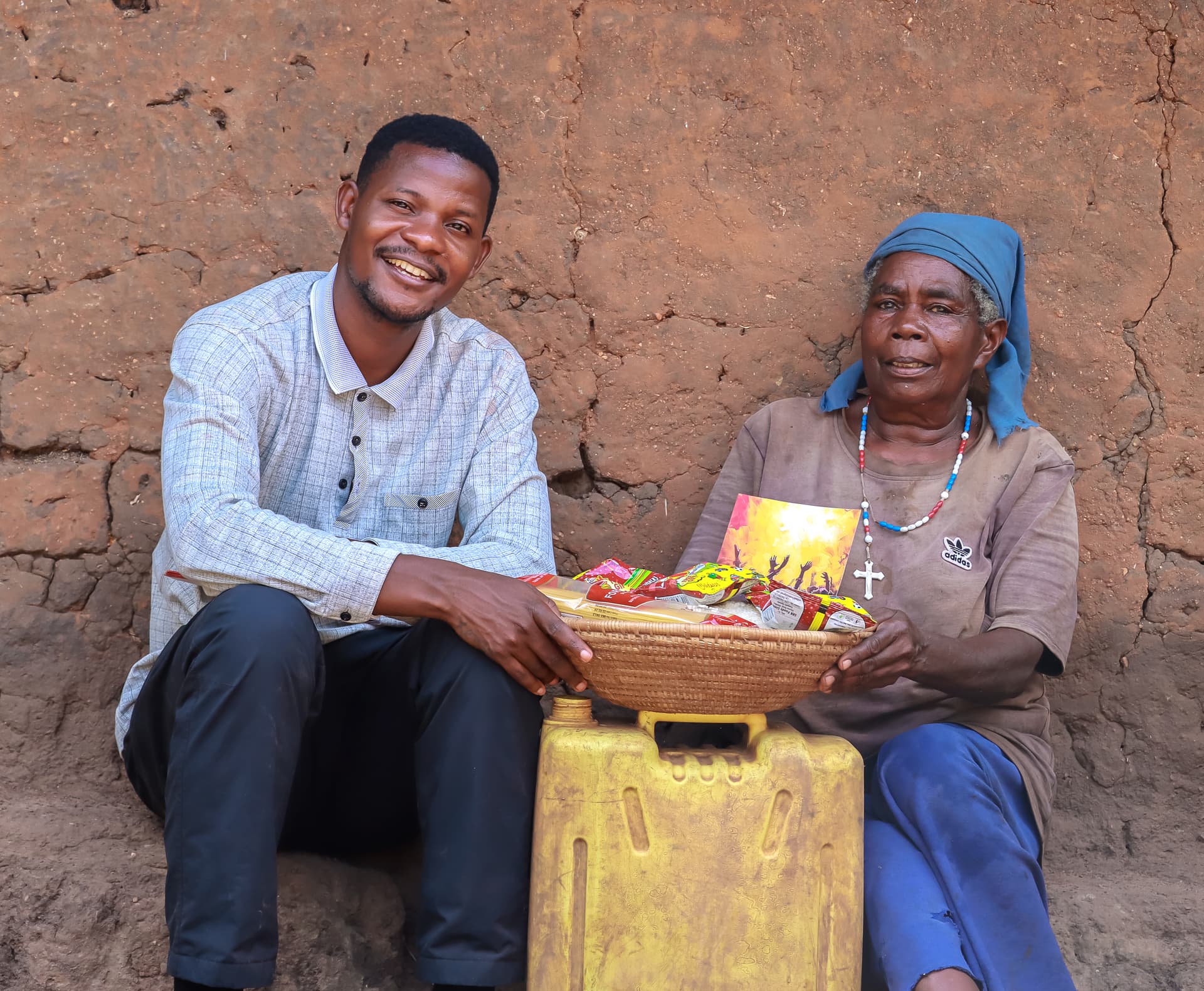 A JFM outreach worker and an elderly woman, Sarah, sit together holding a basket of food gifts against a mud wall.
