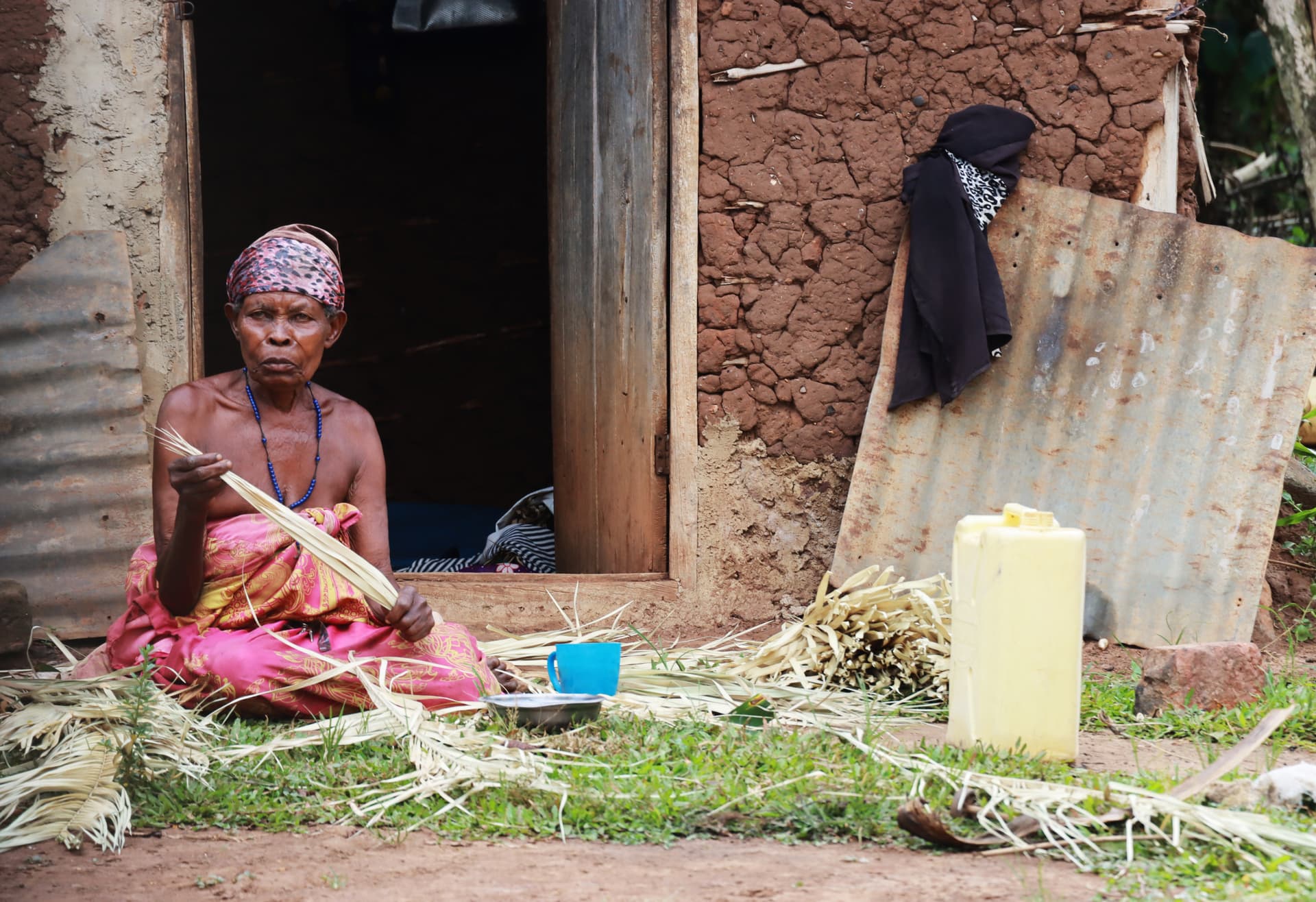 Elder Caroline, wearing a headscarf, sits outdoors preparing mat leaves near her home.