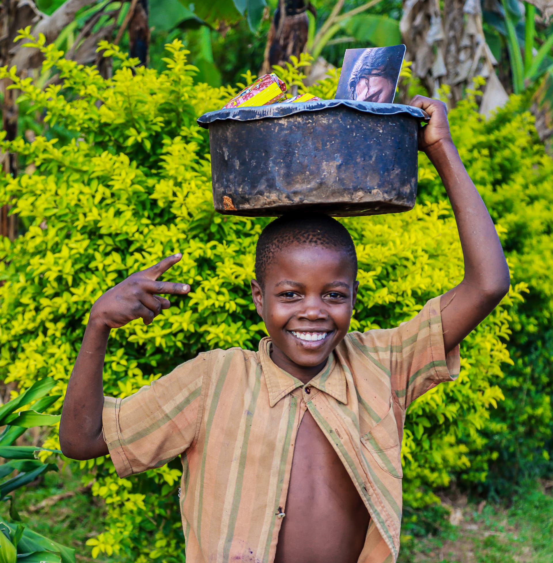 A smiling boy in Kalagala village balances a dark basin on his head while pointing at the camera.