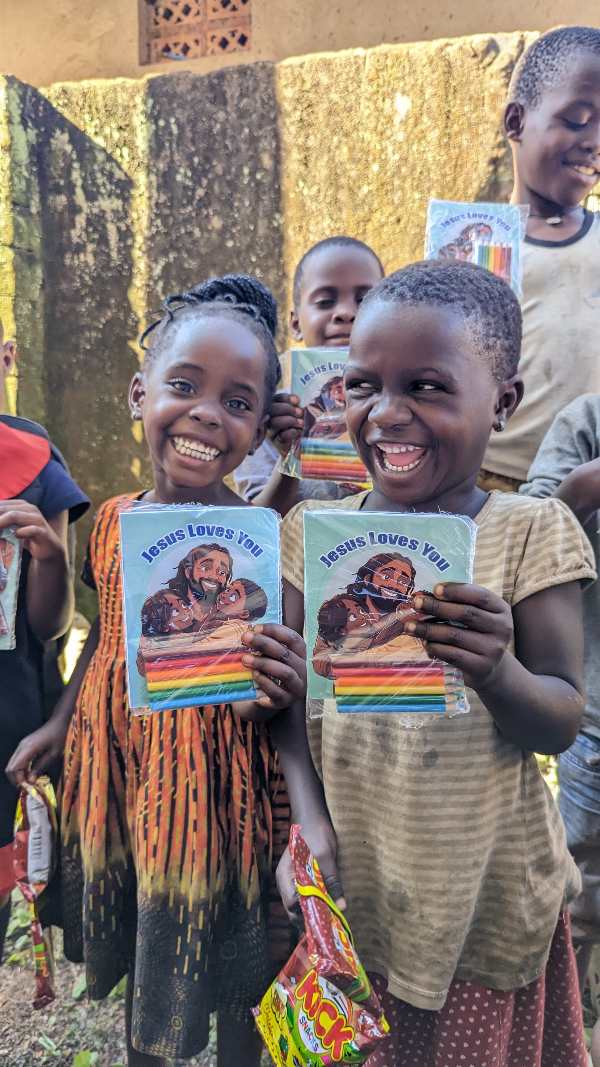 Girls stand outdoors near a brick wall holding colorful children's coloring books.