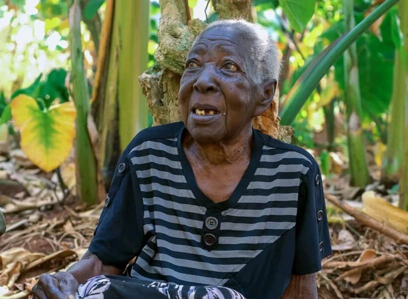 An elderly woman sits outdoors among banana leaves, speaking during a visit.