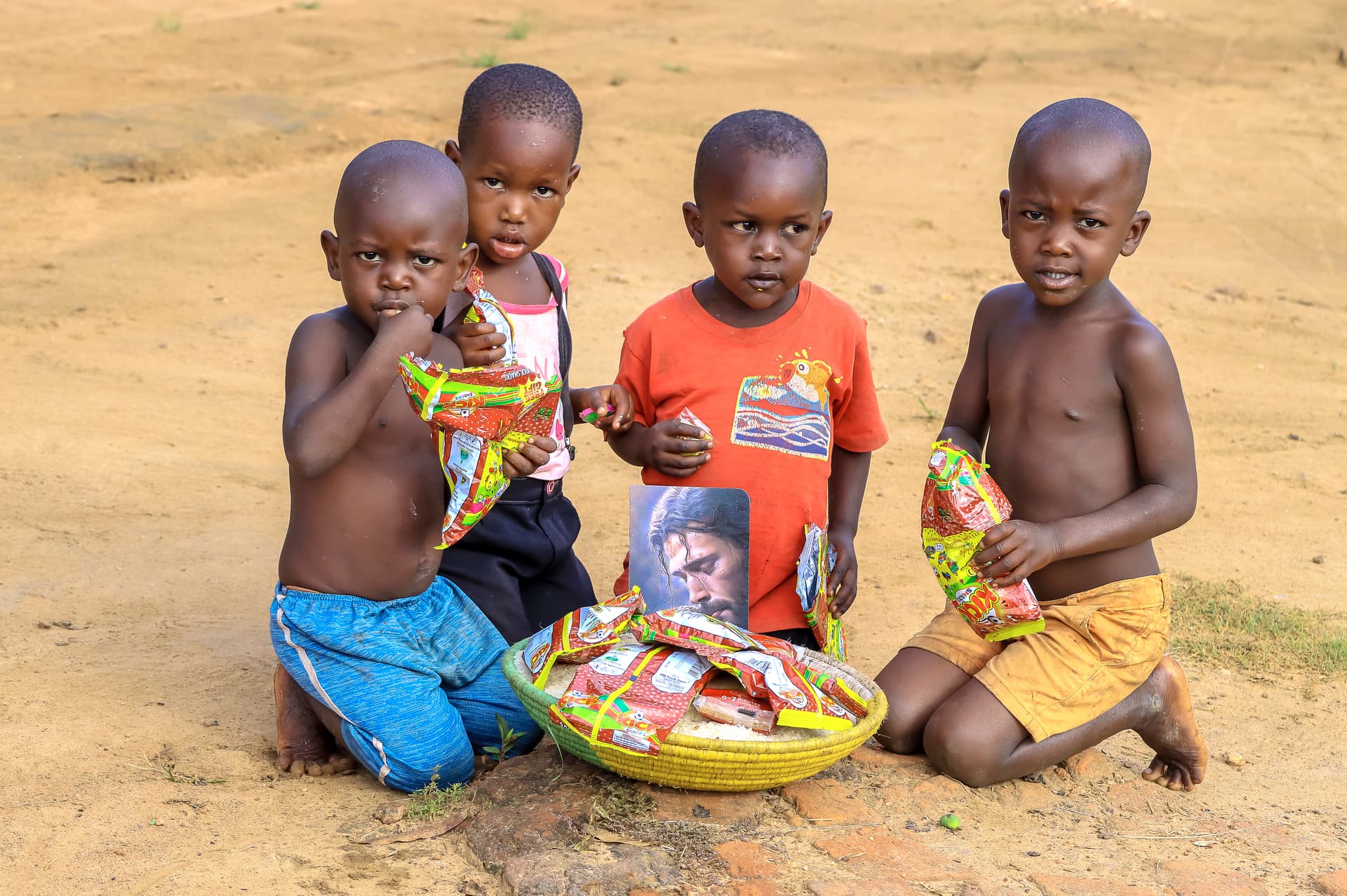 Three young children sitting on the ground around a basin of food packets.