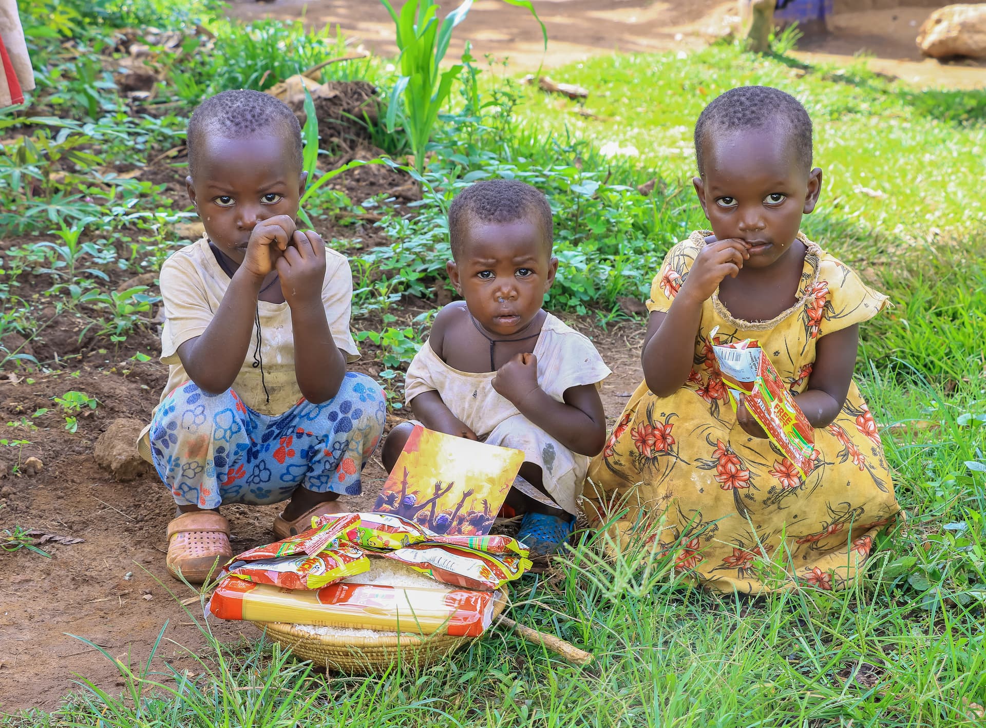 Three children sitting on grass with food packets and gospel tracts.