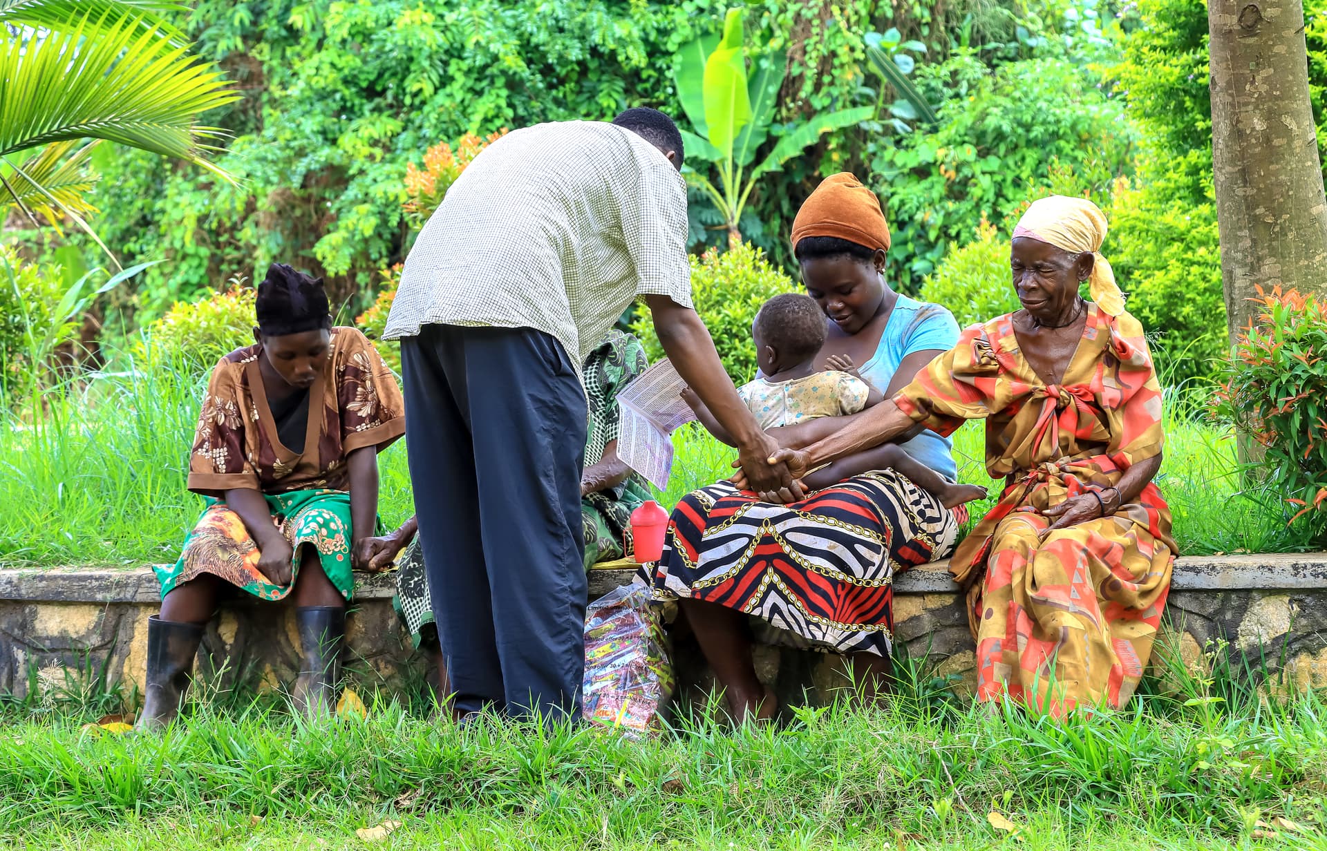 Man in a white shirt hands a gospel tract to a seated group of women and children under trees in Kilembezi village.