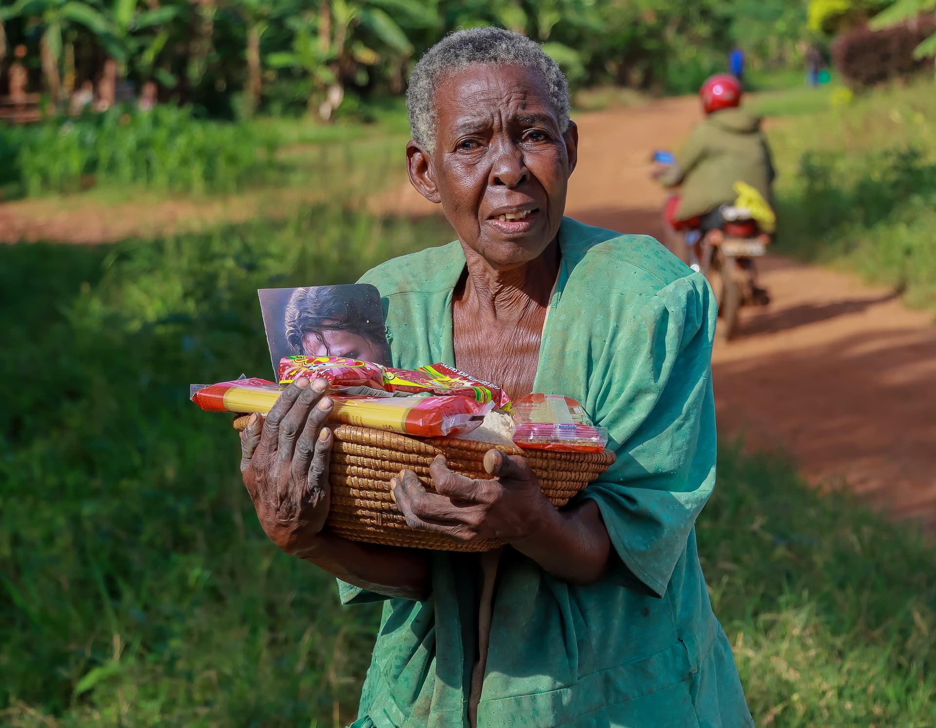 Elderly woman in a teal blouse standing outdoors holding a basket with a gospel tract and snack packets.