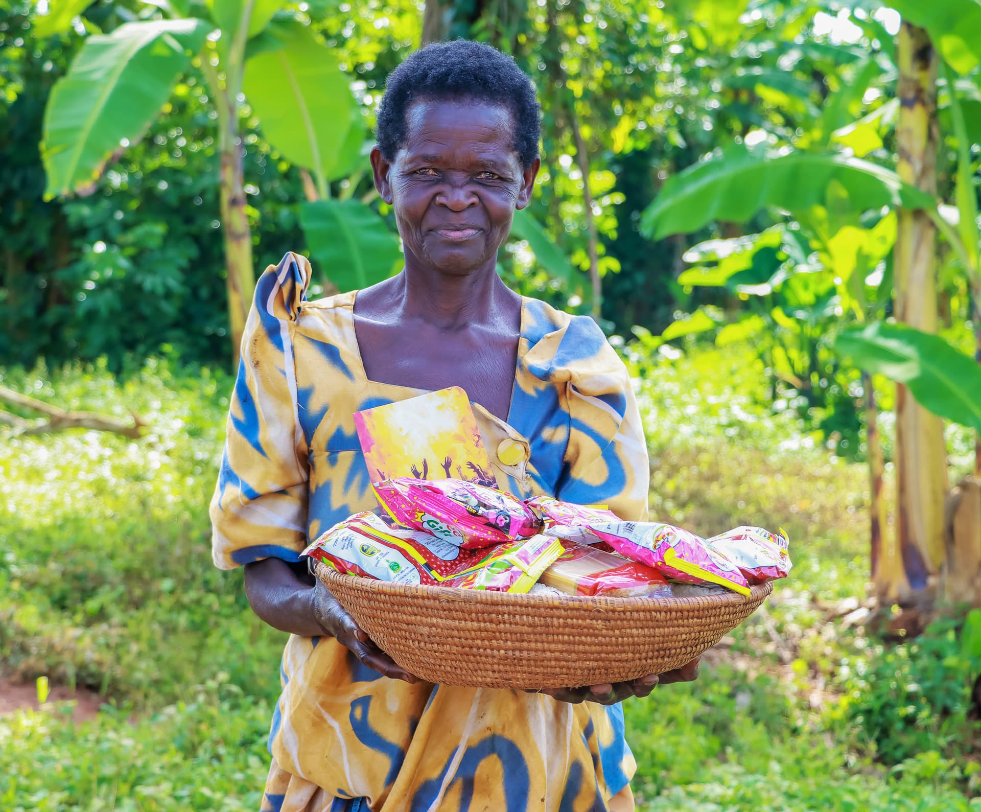 Elderly woman in a yellow and blue patterned dress holding a basket of gospel tracts among banana plants.