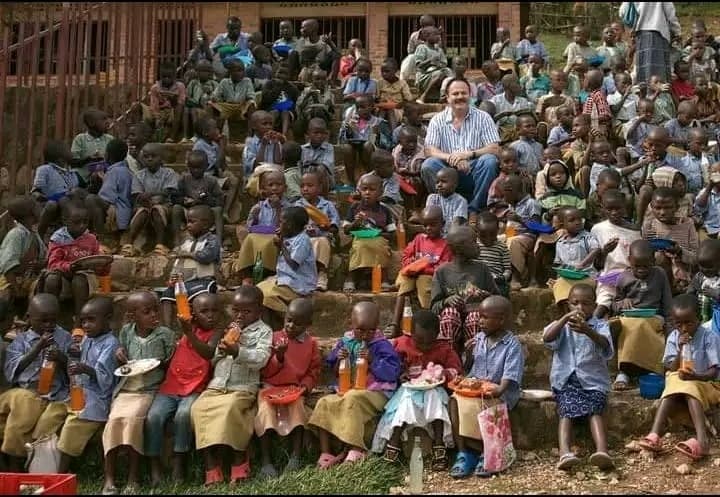 A large crowd of children and adults gathered on benches outdoors at a JFM outreach event, with a brick building in the background.