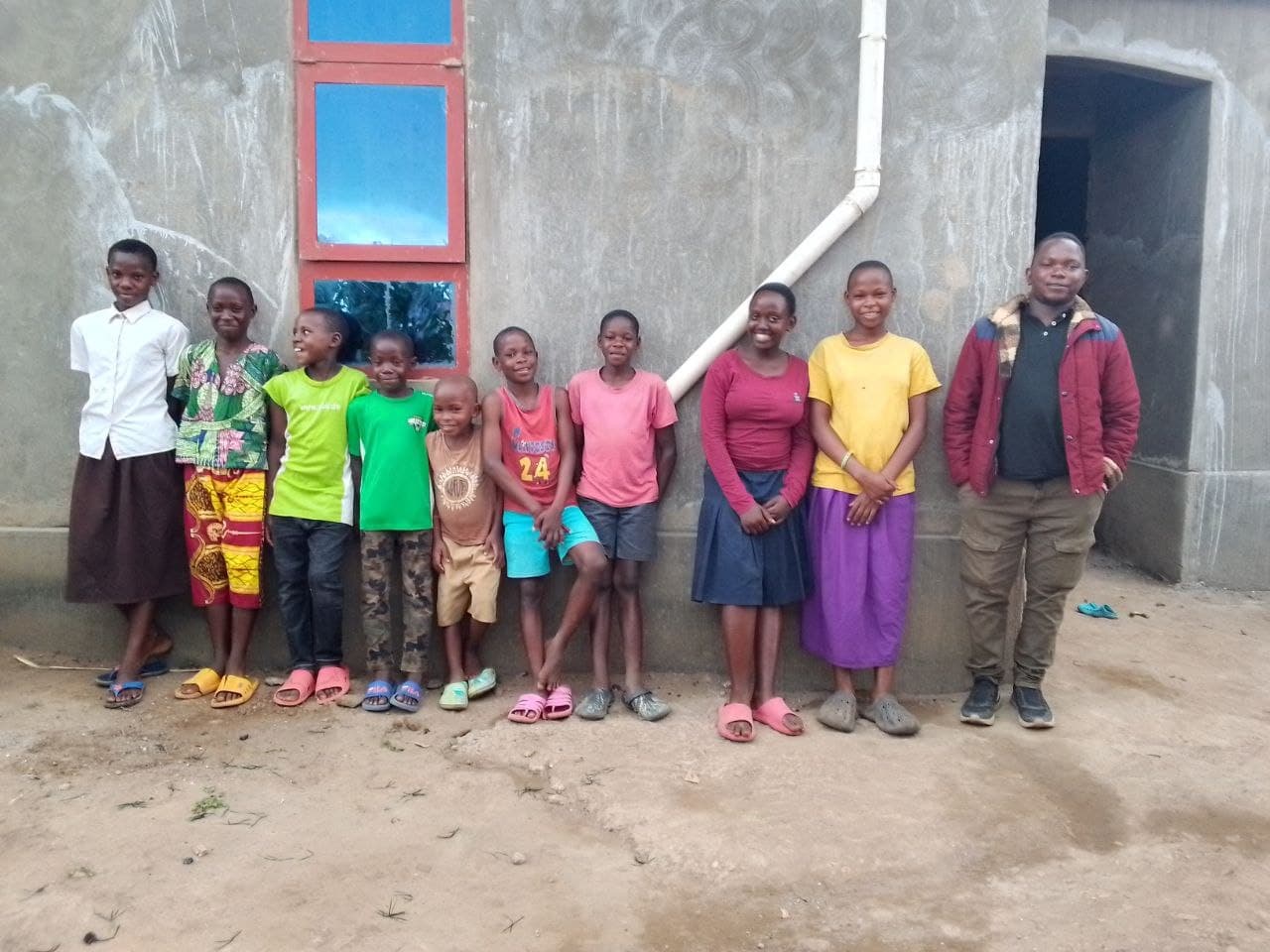 Children from Mashariki Children's Home line up with a JFM staff member in a red shirt against the home's building.