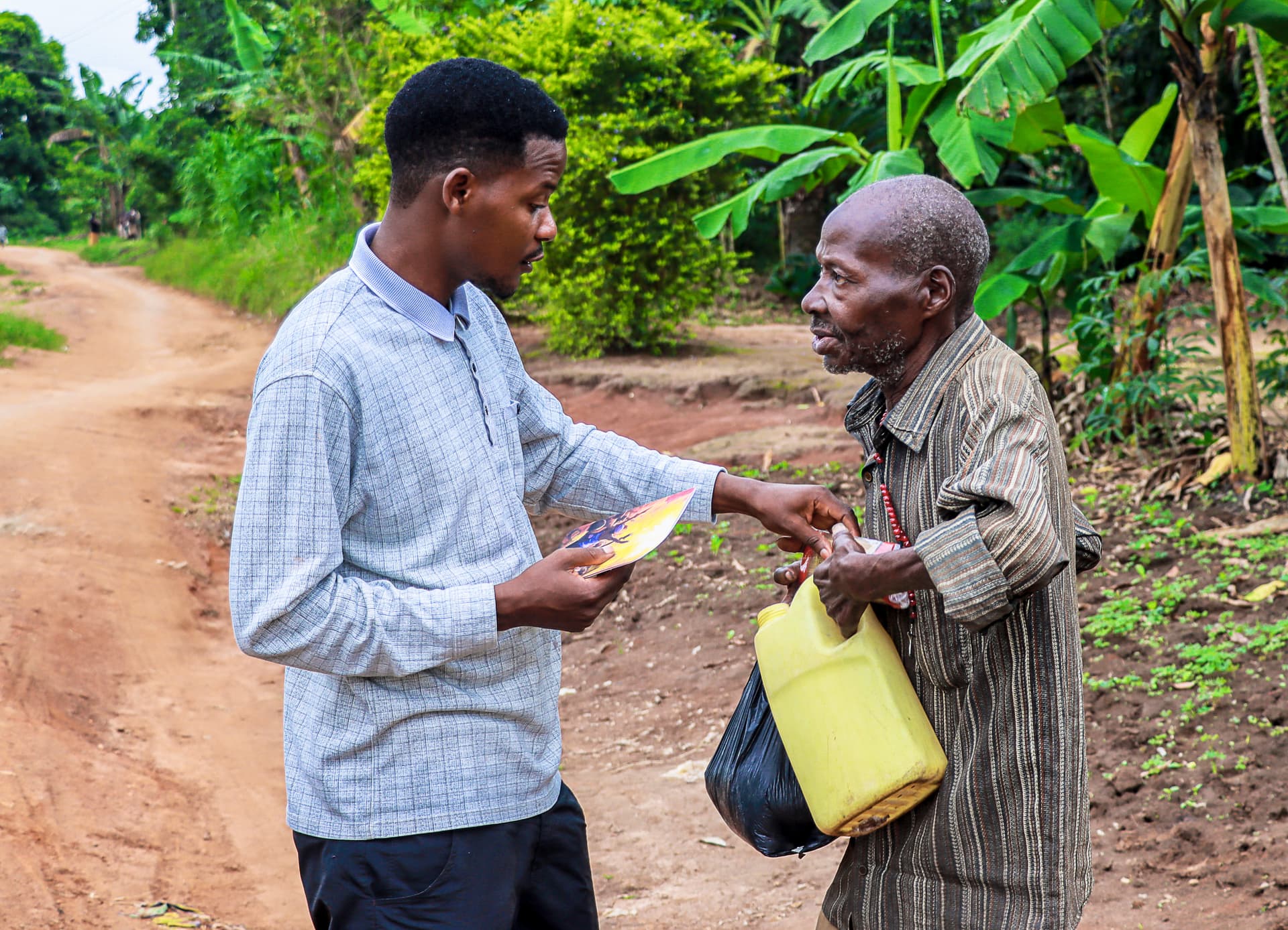 Man in a light blue shirt handing a gospel tract to an elderly man on a rural dirt road.