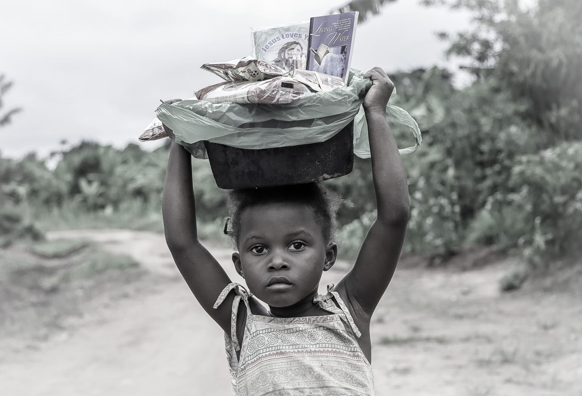 Black-and-white photo of a child balancing a basin overhead with raised arms while walking a dirt road.