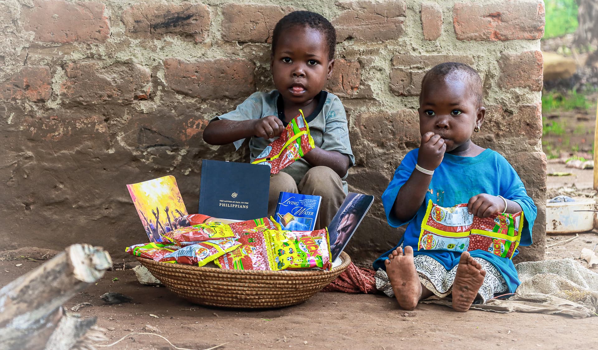 Two toddlers sit against a brick wall eating food packets, with a woven basket of food packets in front of them.