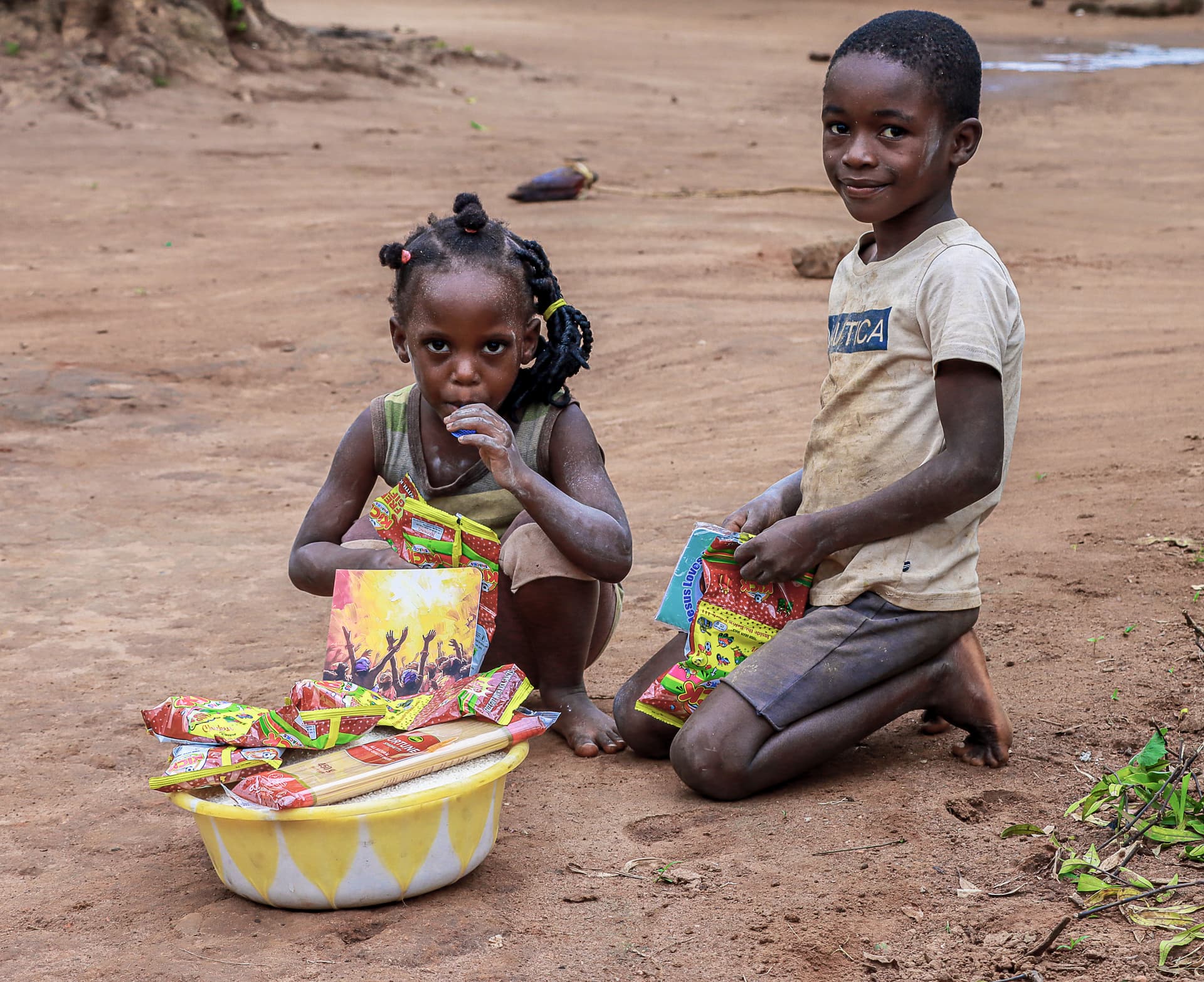 Girl and boy kneeling on dirt ground beside a yellow basin filled with colorful snack packets.