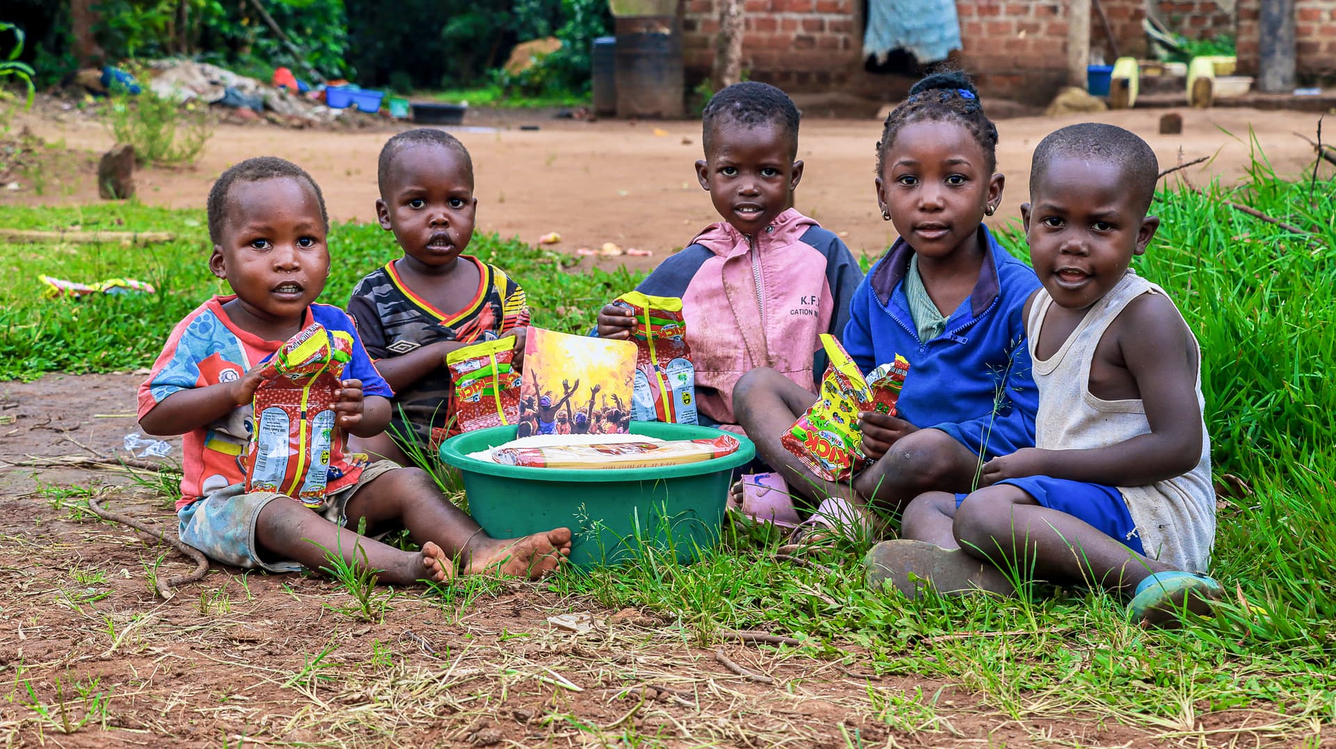 Five young children in colorful clothes sitting on grass around a green basin of food gifts.