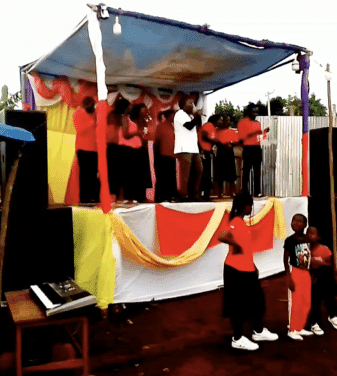 Group of people in red and orange clothing gathered under a decorated outdoor tent at a gospel crusade.