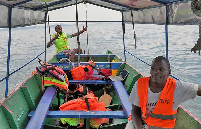 Two men are on a boat on Lake Victoria, one steering and one wearing an orange life vest, with life jackets stored aboard.