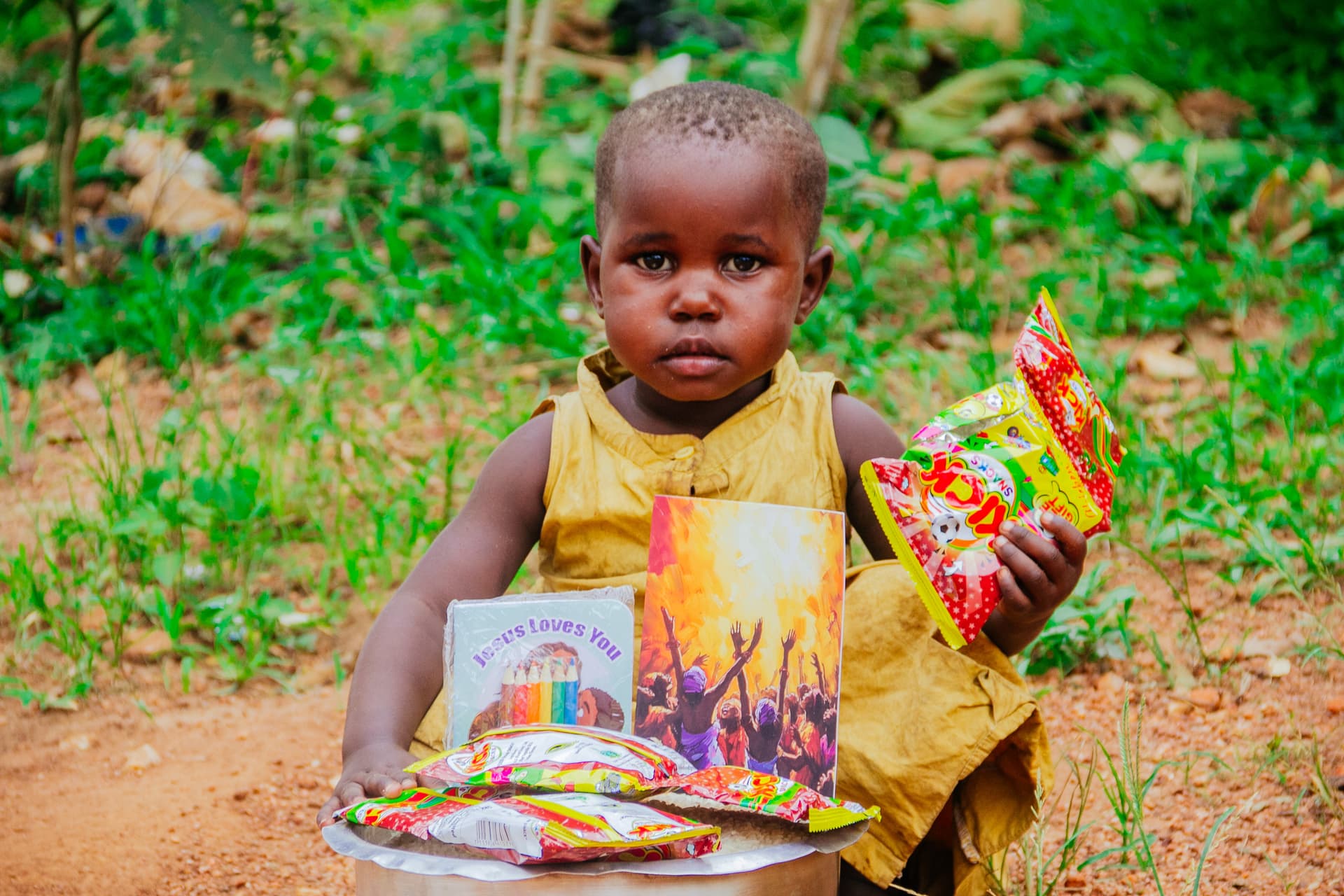 A toddler in a yellow dress sits outdoors holding a food packet, with a plate of food gifts nearby.
