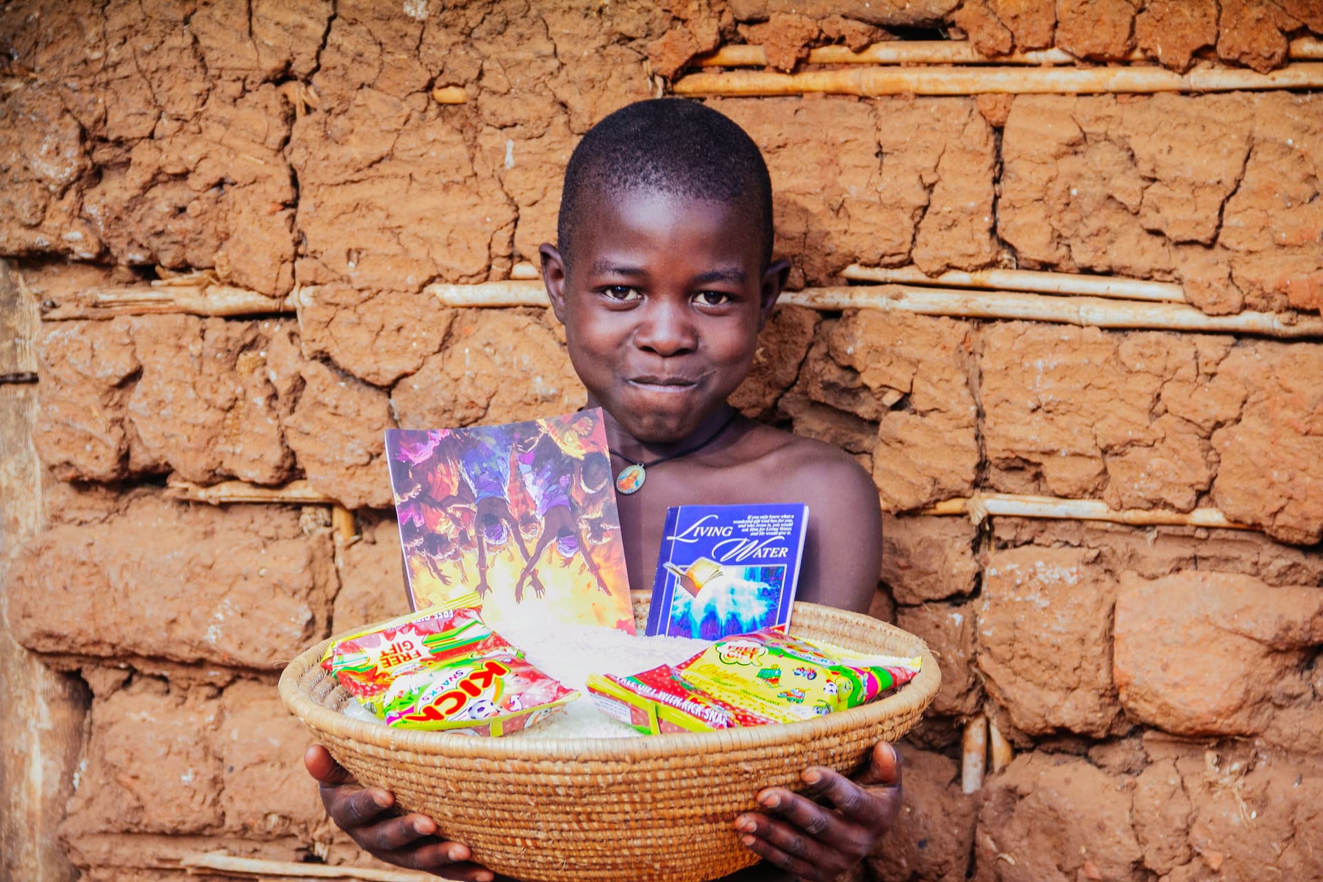 A boy stands against a brick wall holding a woven basket of food packets and a gospel tract.
