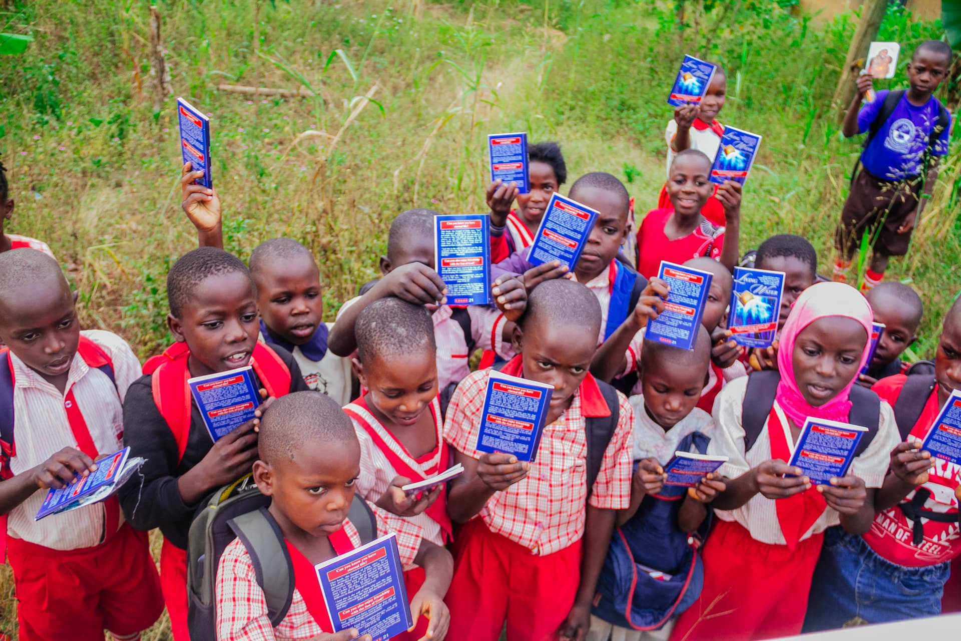 A crowd of students at Kemu Education Centre in red-and-white uniforms hold up blue gospel booklets outdoors.