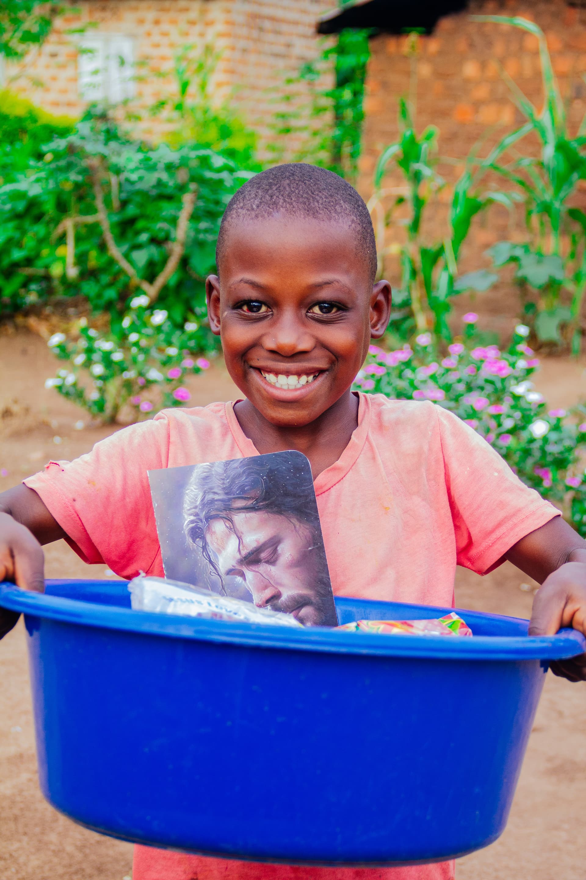 A boy in a salmon-colored shirt smiles while holding a blue basin filled with food packets.