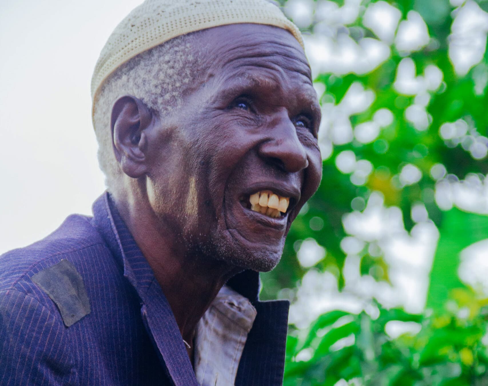 An elderly man in a knit cap and dark jacket smiles outdoors near greenery.
