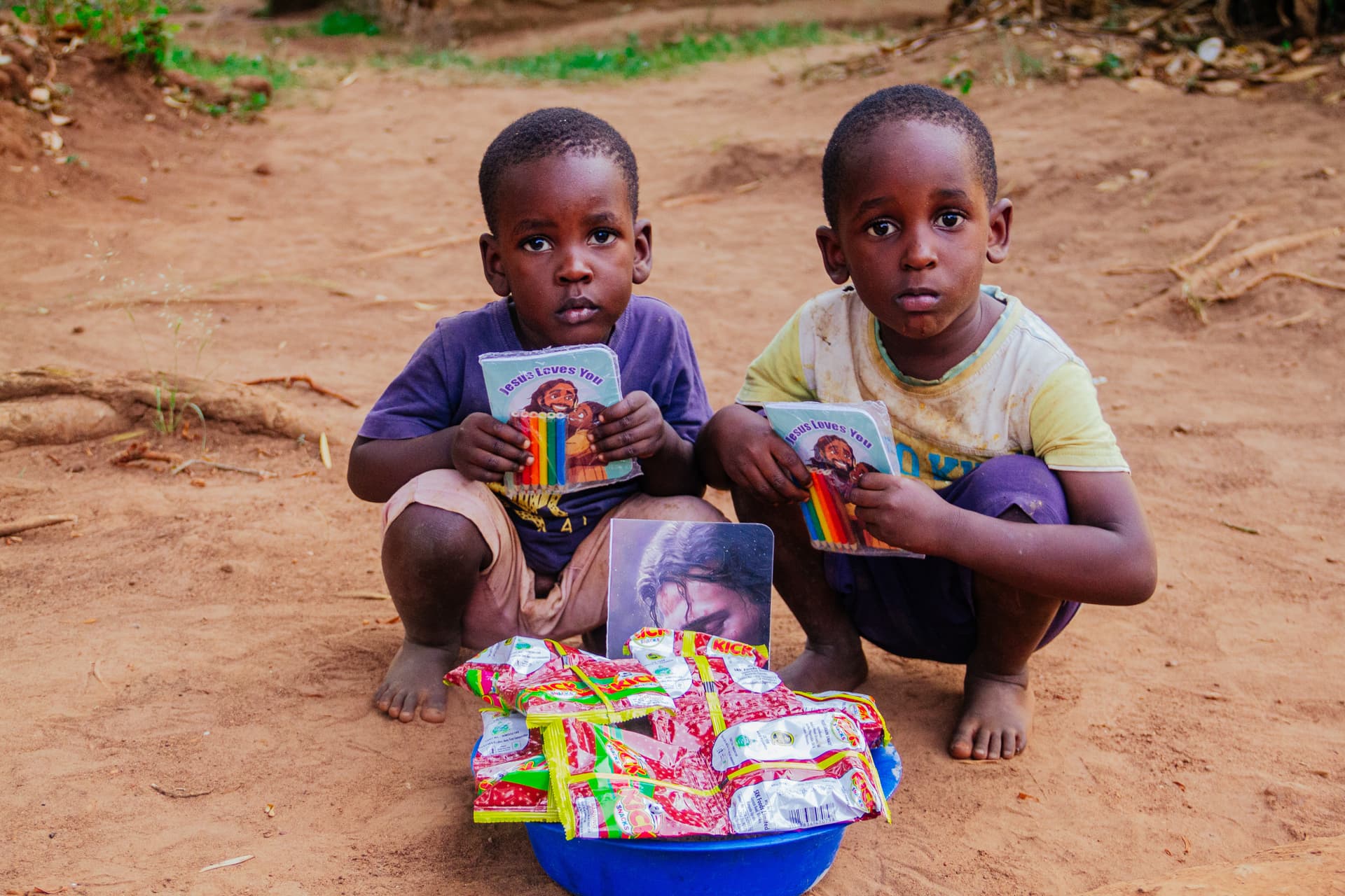 Two boys crouch together on a dirt path, each holding food packets beside a blue basin of supplies.