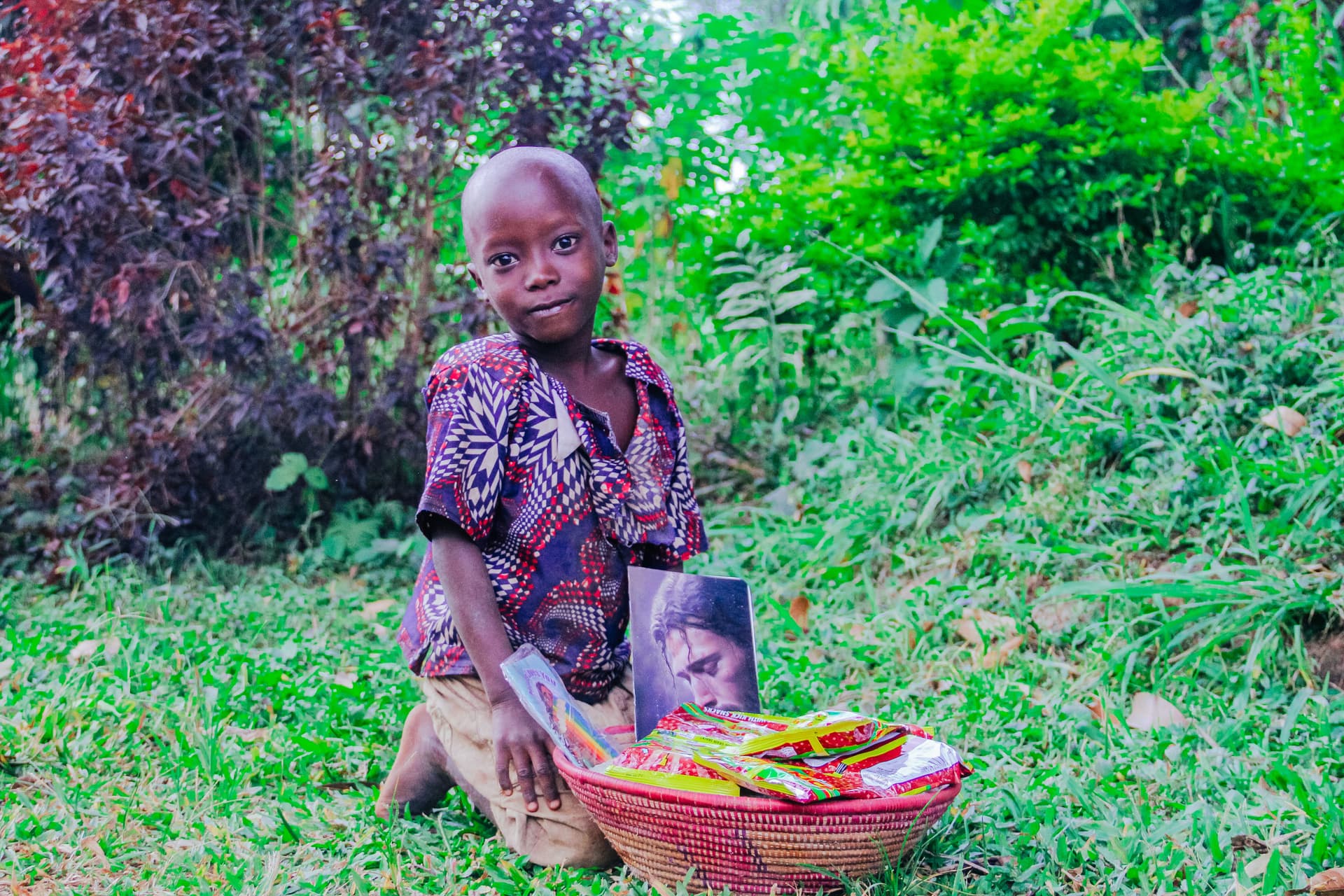 A girl kneels in the grass smiling, holding a red basin filled with food packets and a gospel tract.