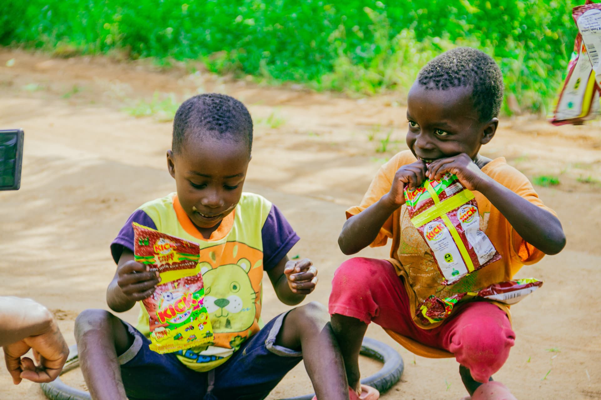 Two boys sitting on the ground eating snack packets outdoors
