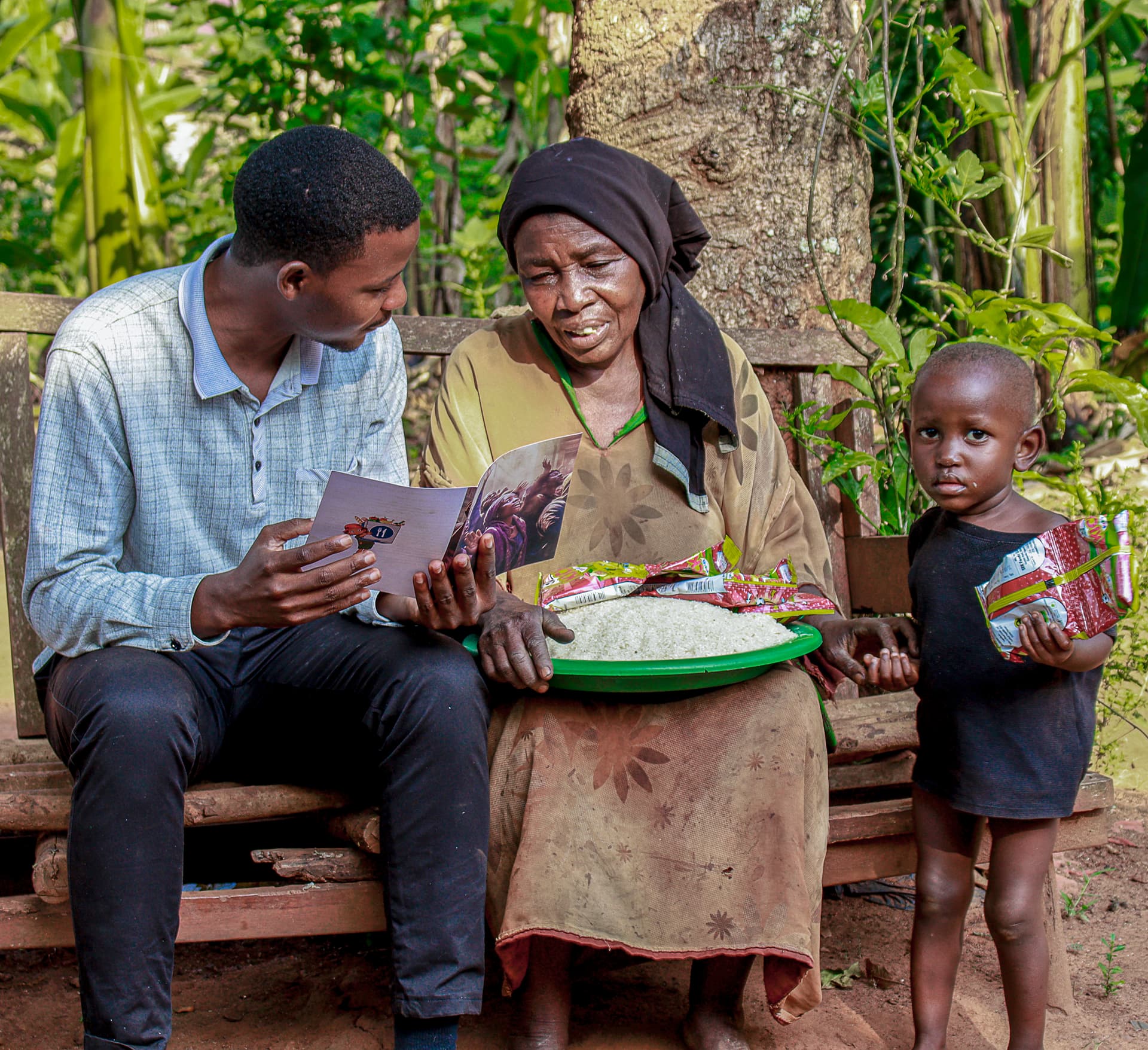 A man reads a gospel booklet with a woman and a young boy seated on a wooden bench outdoors, a basin of food packets at their feet.