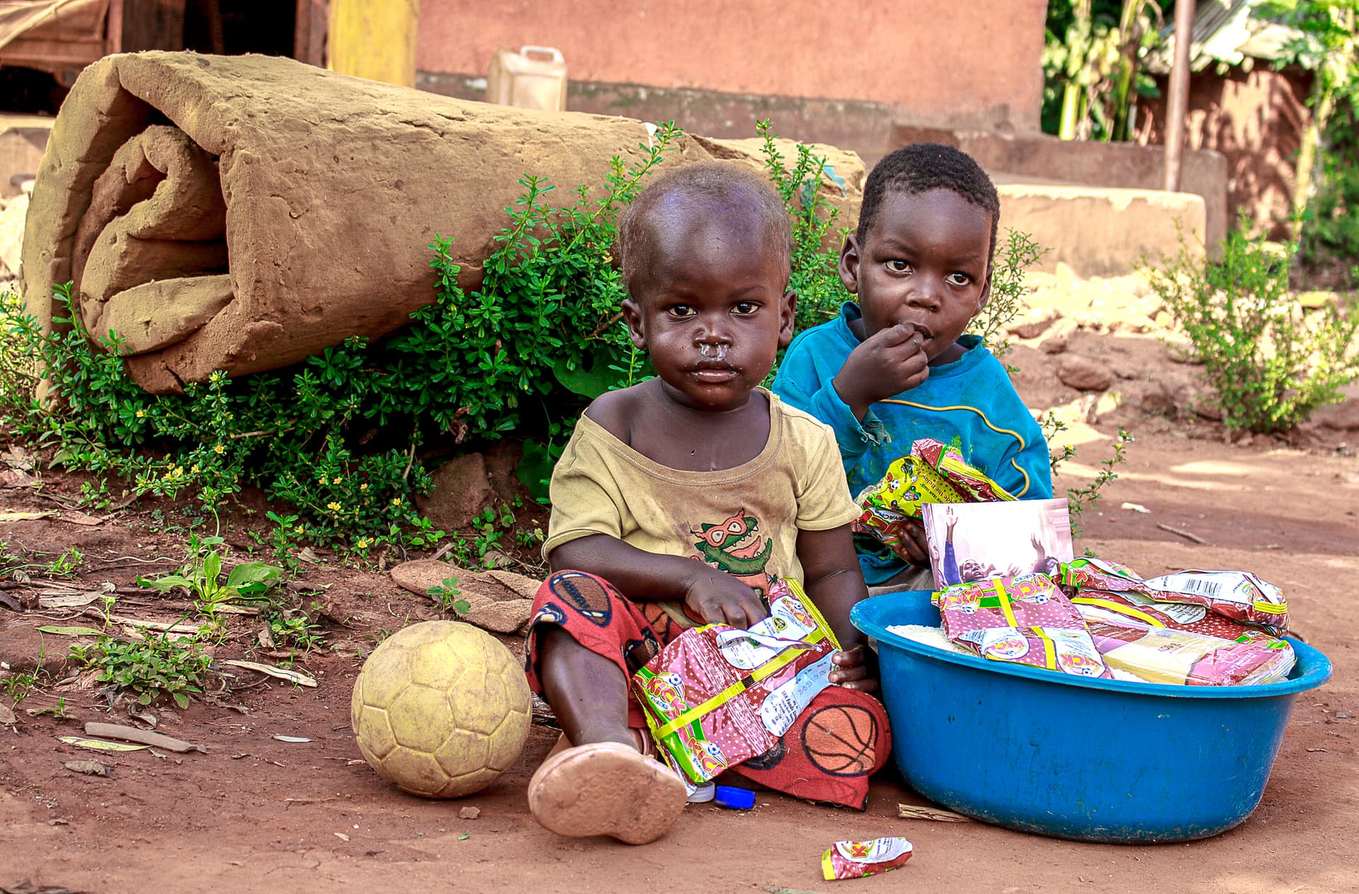 Two young children sitting outdoors next to blue basins of food and a soccer ball