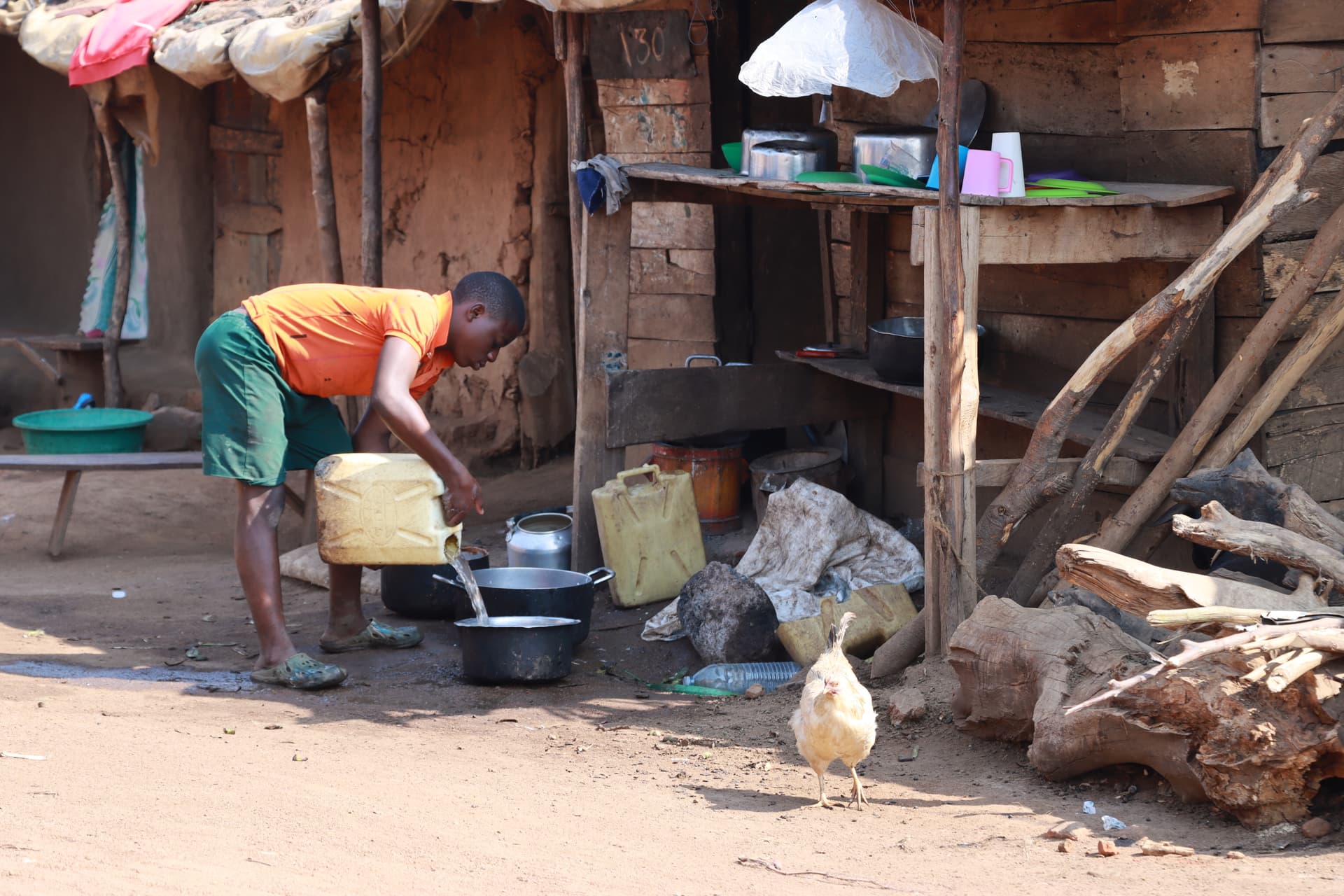 A child cooks at an outdoor kitchen station beside a mud house.