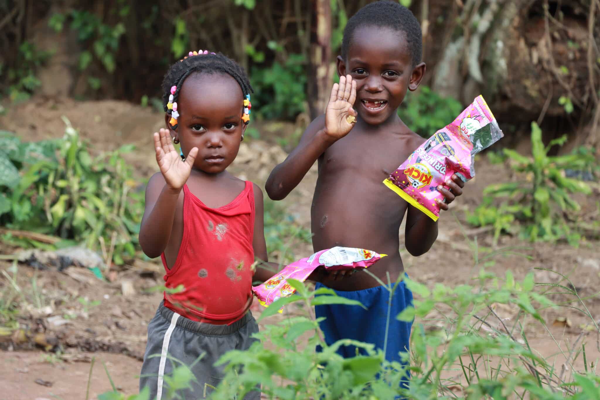 Two boys outdoors holding snack packets, one waving at the camera