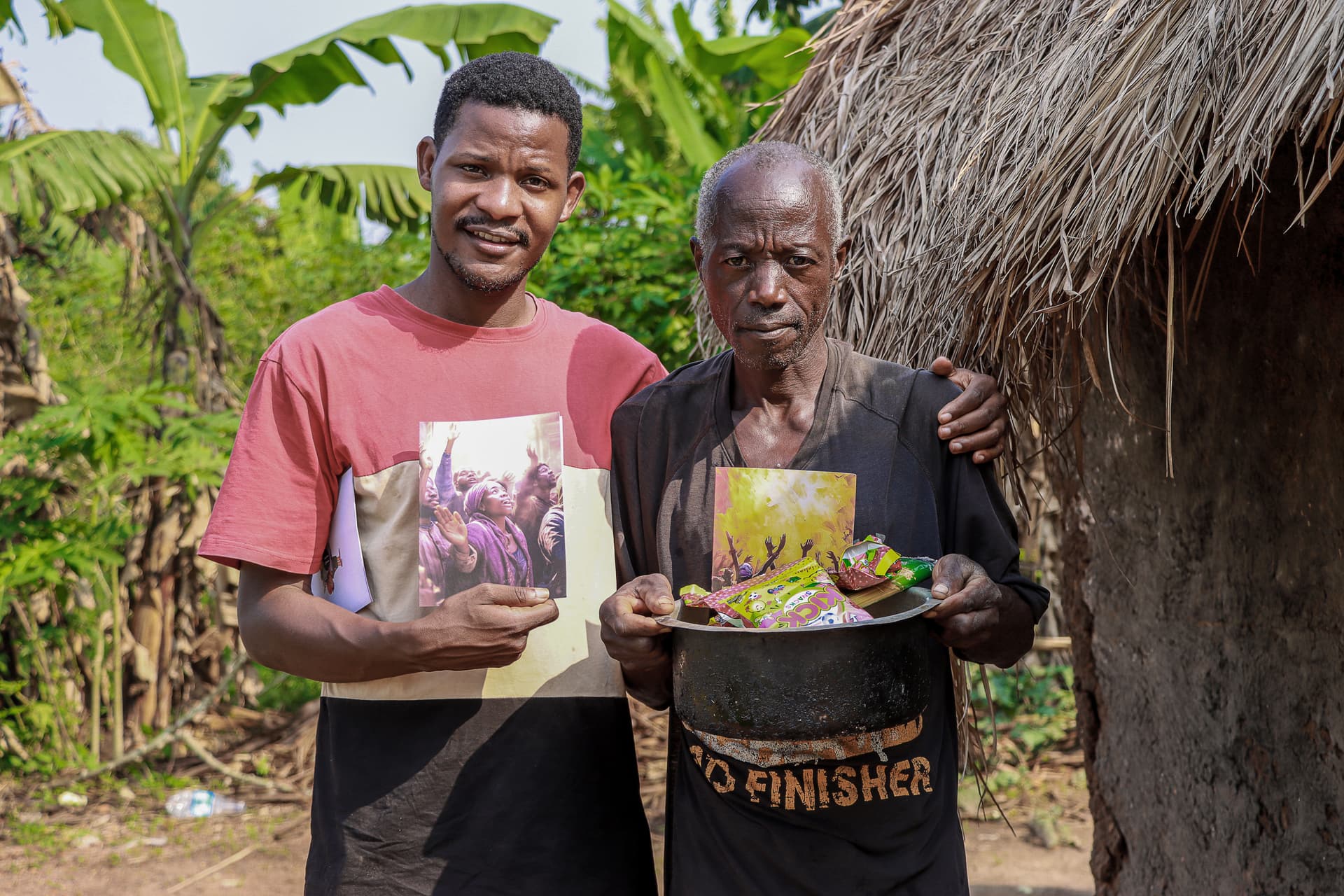 Two men, one holding a gospel tract and bowl of fruit, standing with an older man outside a grass hut.